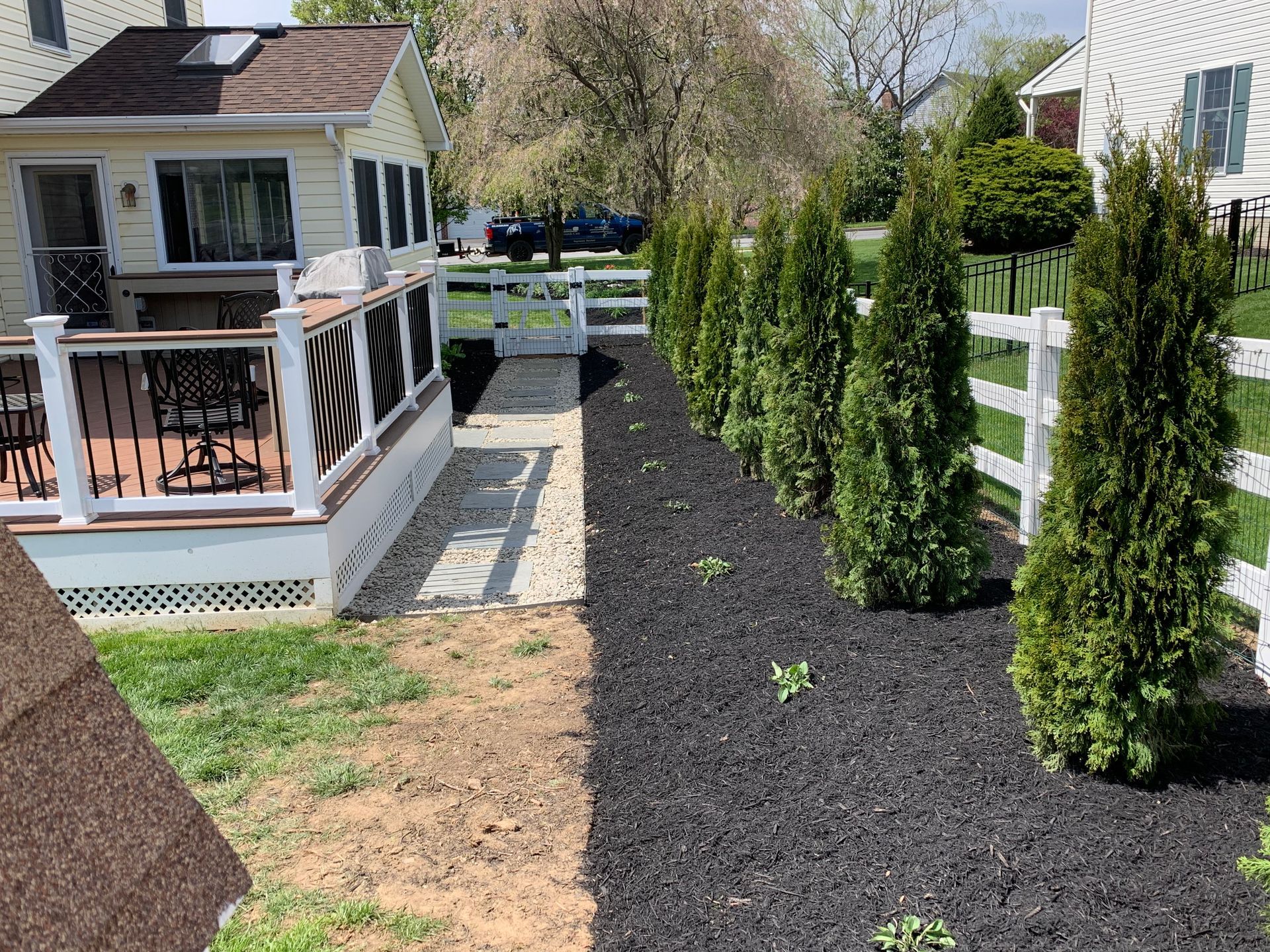 A side yard features a deck with black railings, a stone walkway, black mulch, and a row of evergreen trees by a fence.