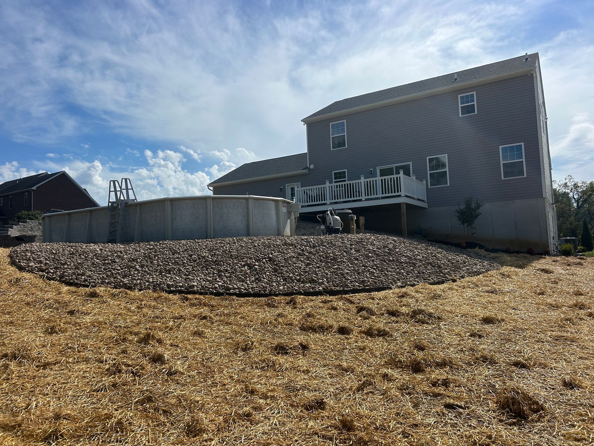 A grey two-story house with a deck next to an above-ground swimming pool, surrounded by a rock bed and a grassy yard.