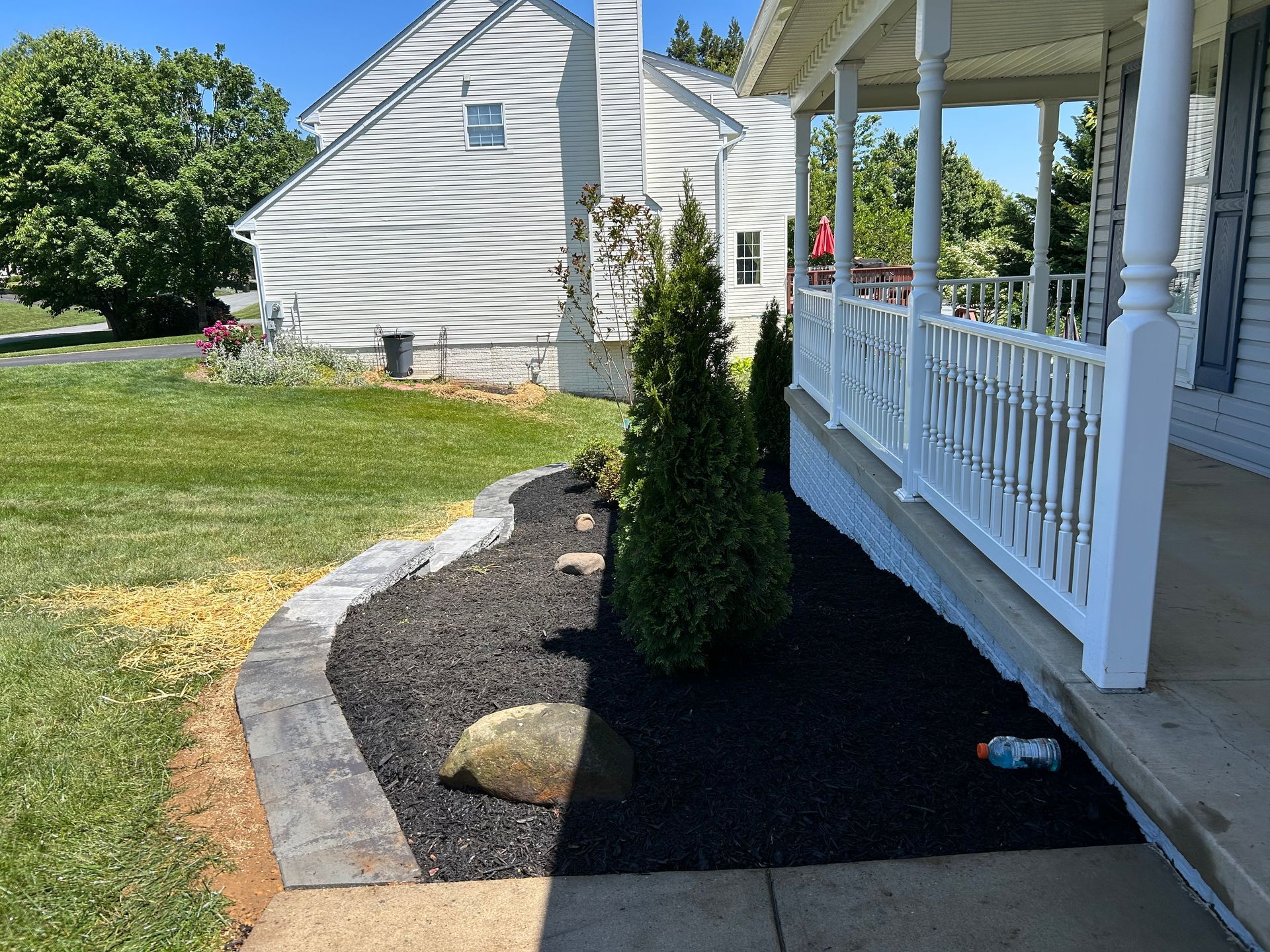 A landscaping bed with black mulch, a stone border, and evergreen shrubs running along a white house porch.