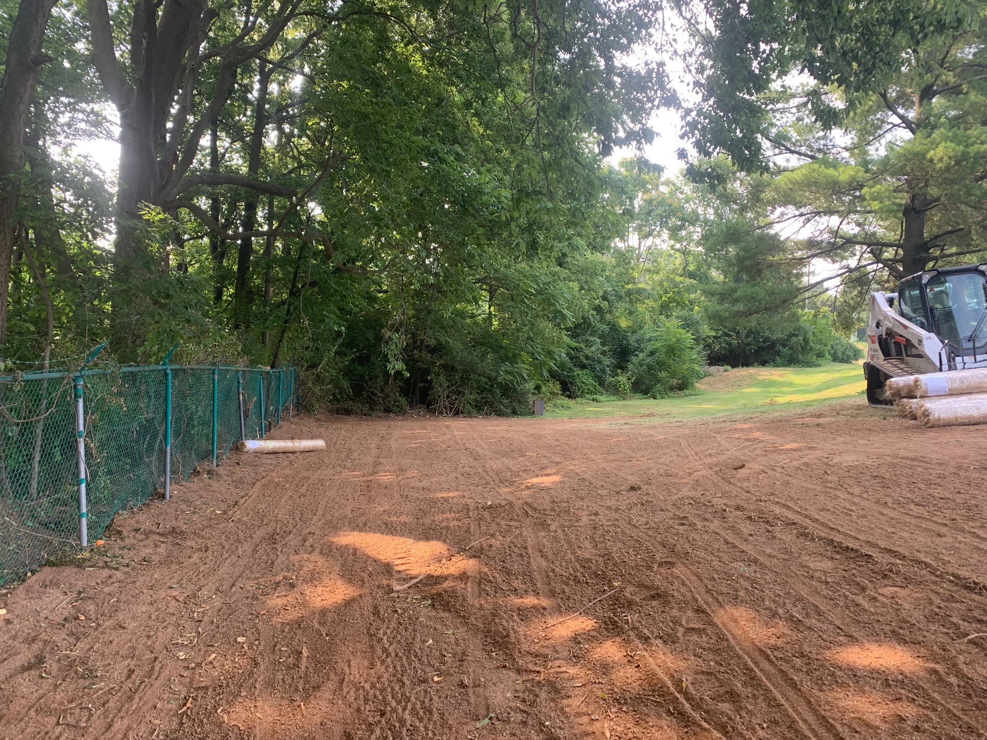 A cleared, dirt ground area adjacent to a green chain-link fence, with trees in the background and a skid steer on the right.