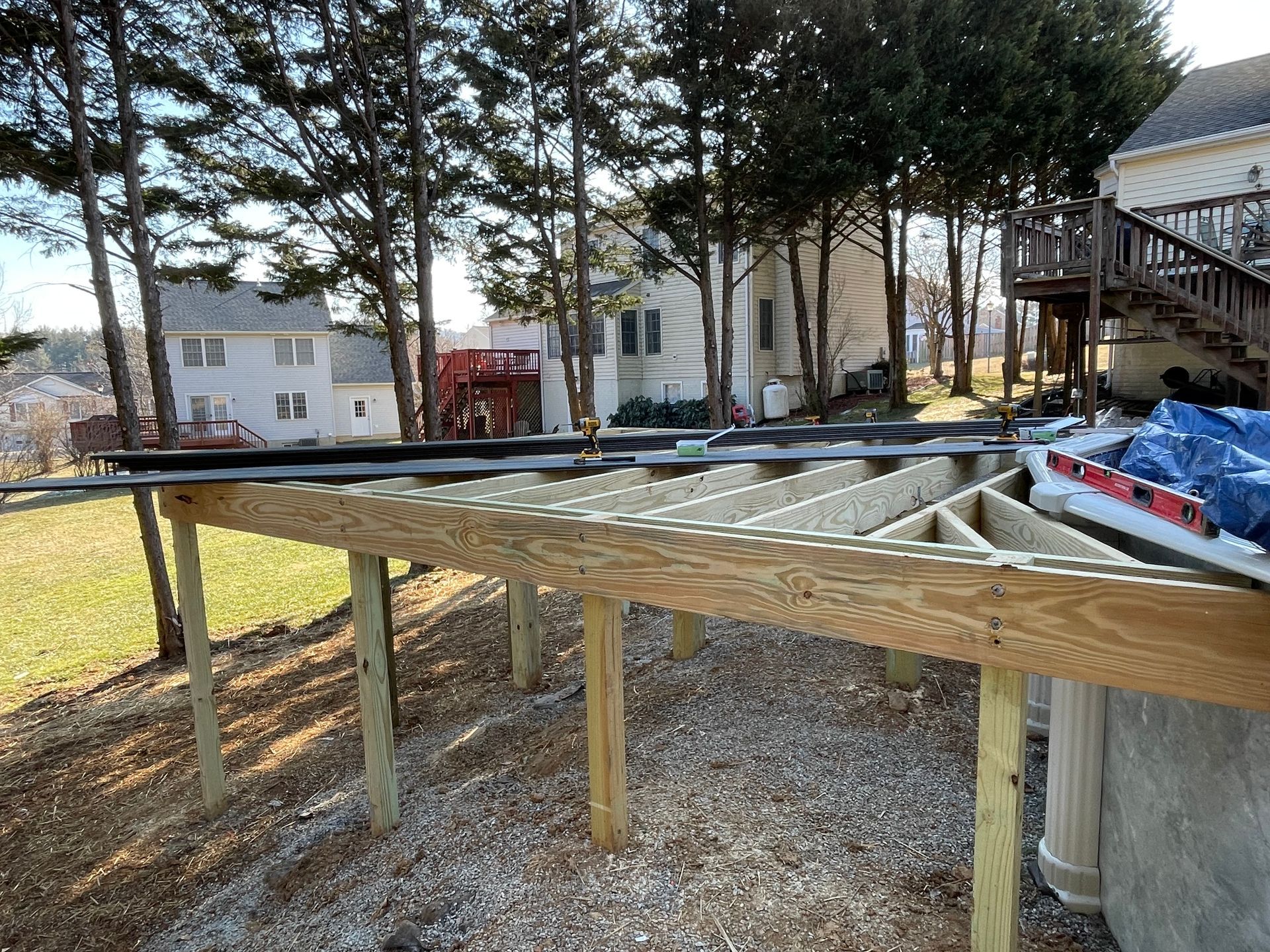 A wooden deck frame under construction in a residential backyard, featuring a partial floor layout and support posts.