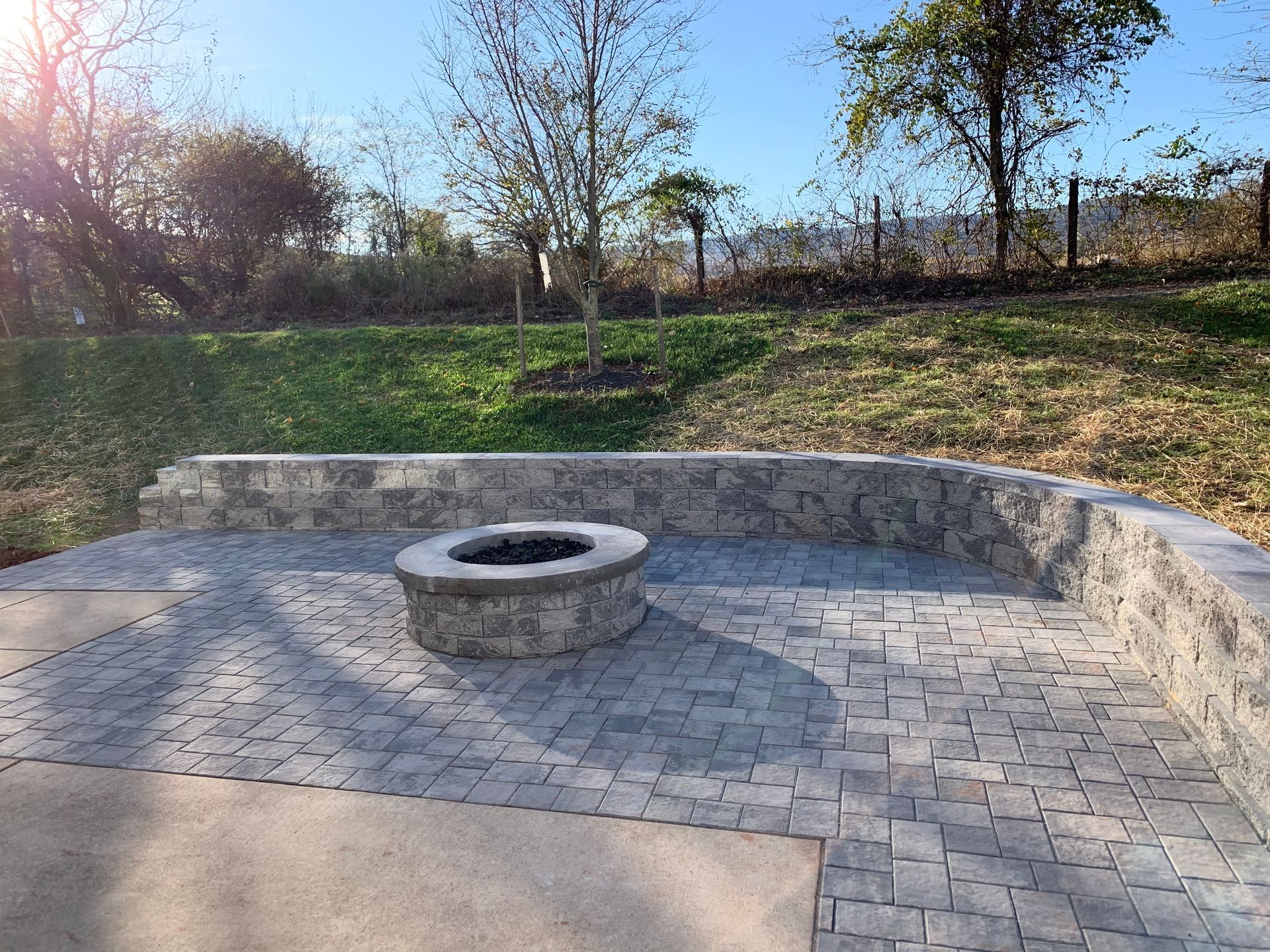 A circular stone fire pit sits on a grey paver patio, backed by a curved stone retaining wall in a grassy backyard.