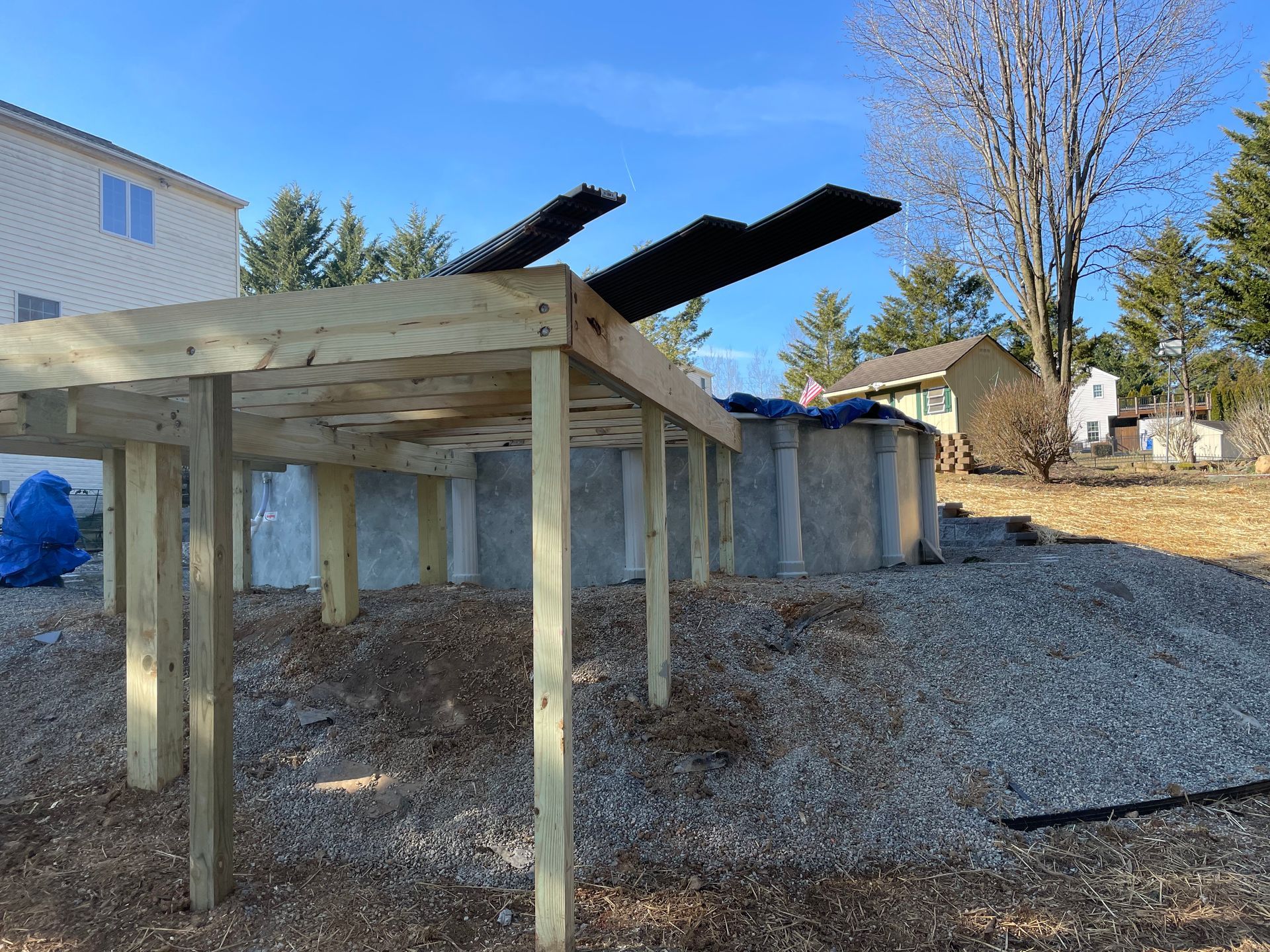 A wooden deck frame is being constructed on a sloped, gravel-covered yard next to an above-ground swimming pool.