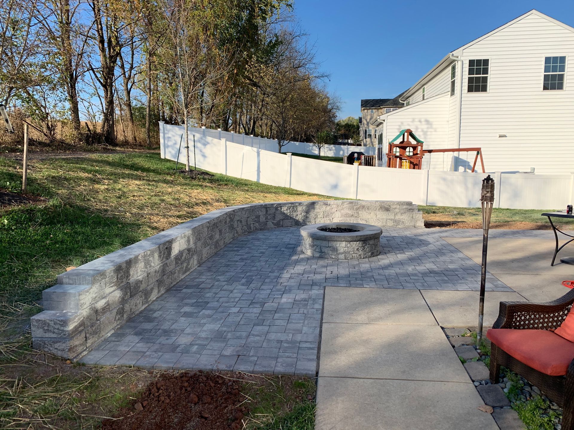 A backyard patio featuring a stone retaining wall, a circular fire pit, and stone pavers near a white house and fence.
