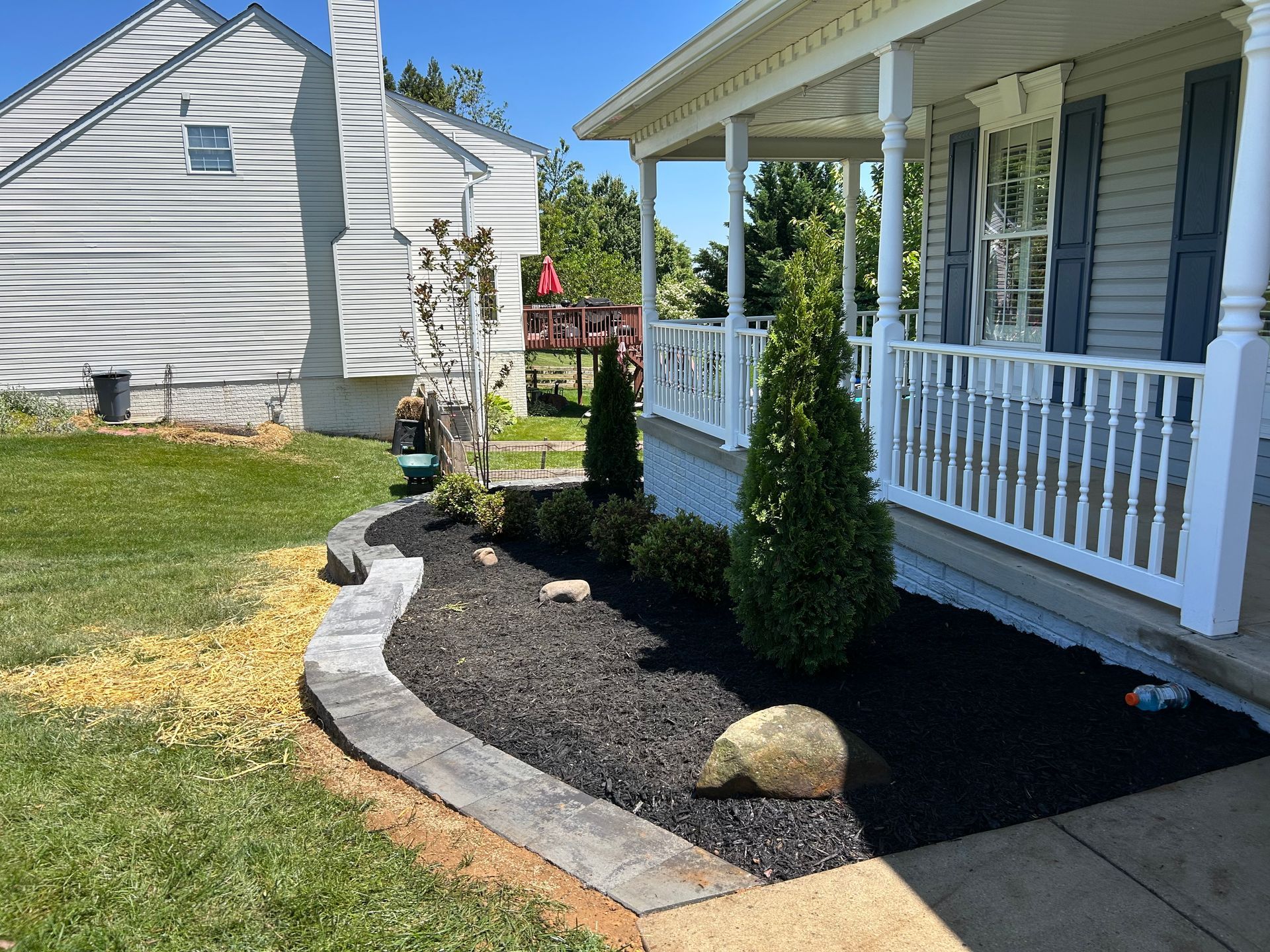 A landscaped flower bed with black mulch, a stone border, and small shrubs next to the porch of a white house.