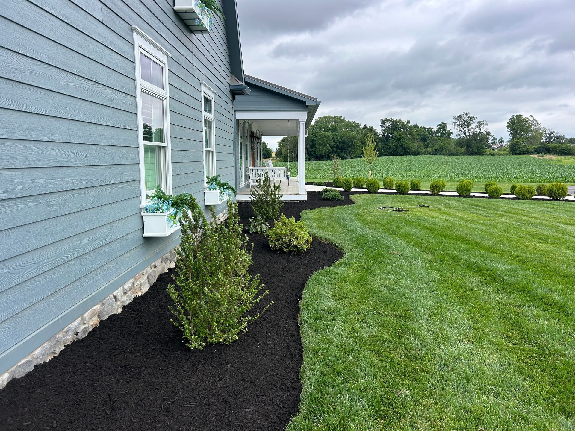A light blue house with a covered porch and fresh dark mulch landscaping along the front, beside a green grassy yard.
