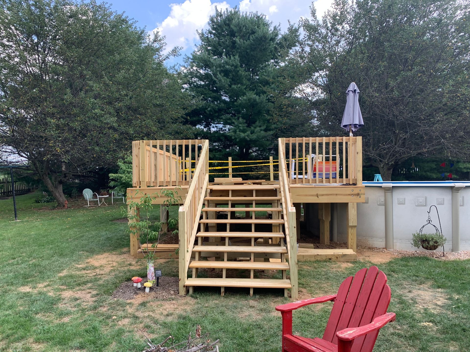 A wooden deck with a central staircase leads to an above-ground pool in a grassy backyard with a red chair in front.
