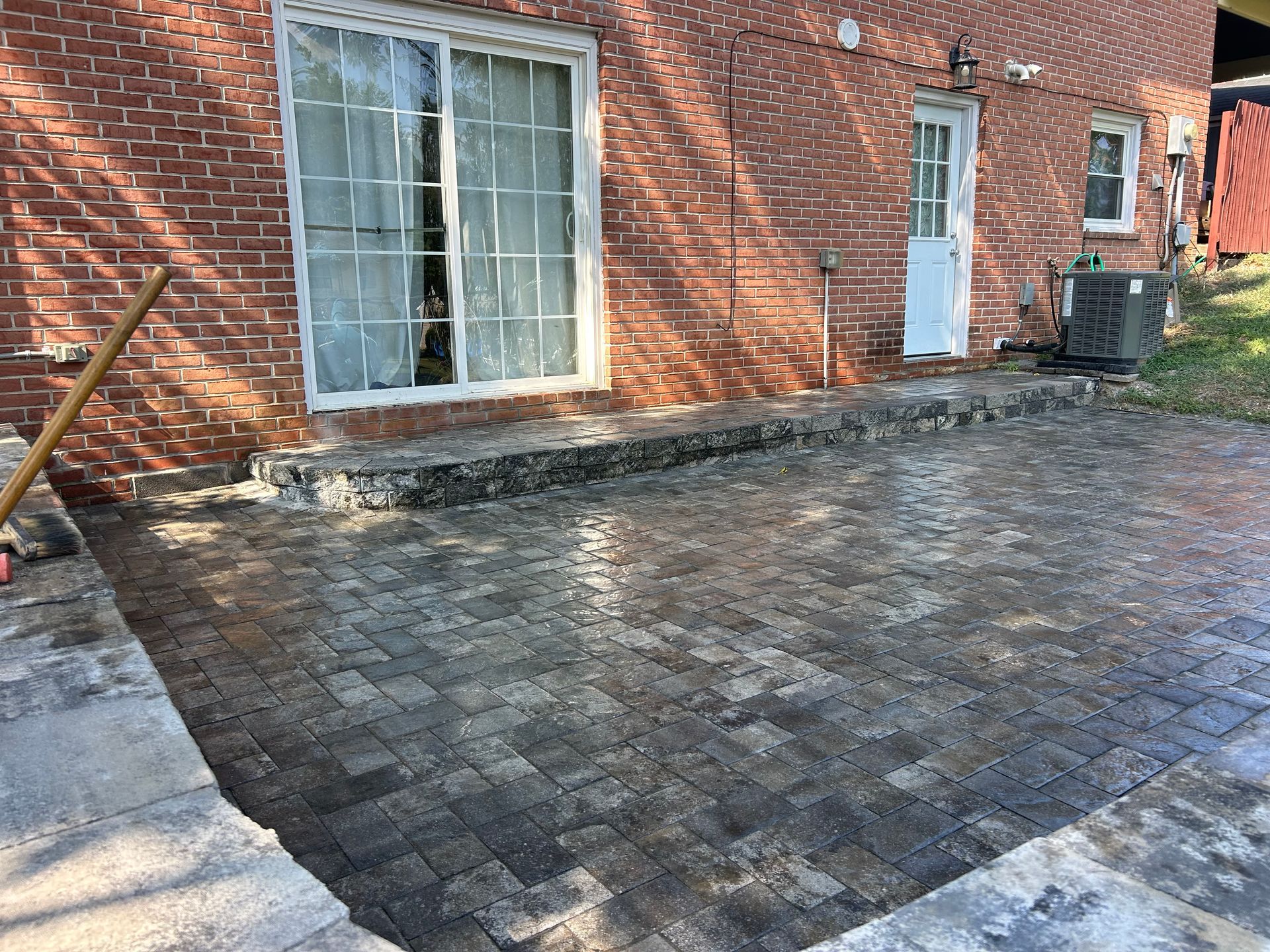 A newly installed stone paver patio sits against a brick house with sliding glass doors and a white door.