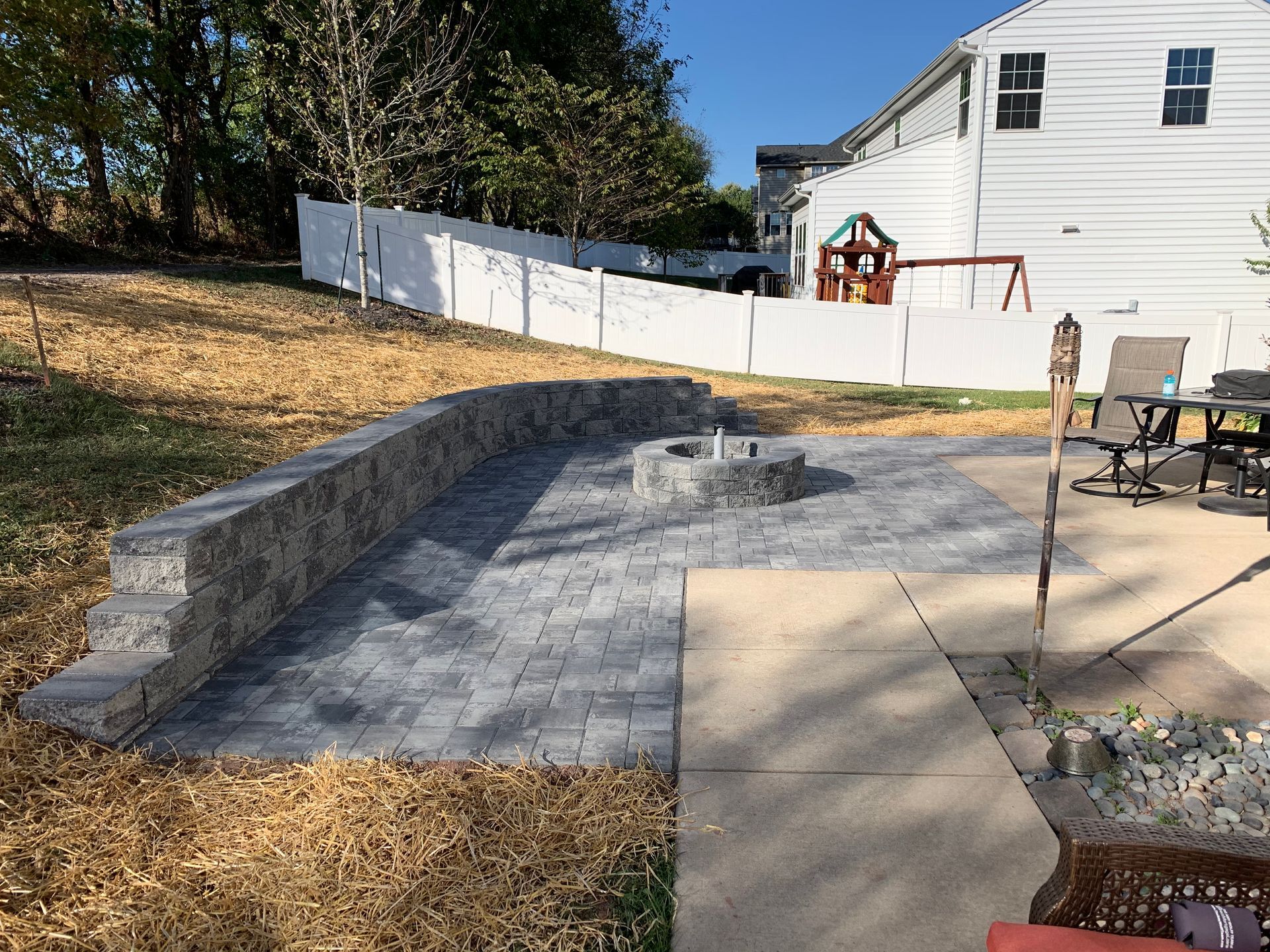 A gray stone patio featuring a circular fire pit and a curved retaining wall in a backyard with a white fence.