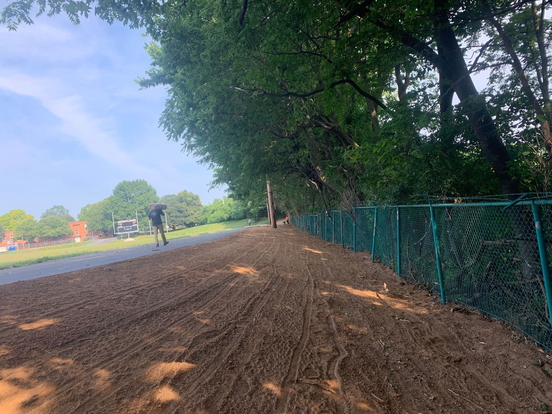 A dirt path runs alongside a green chain-link fence and a line of trees, with a person walking in the distance.
