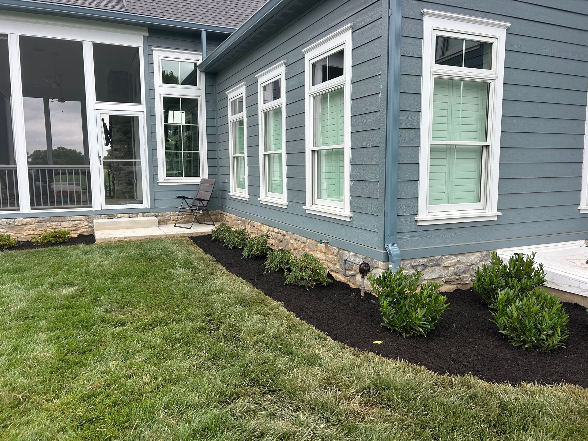 A blue-sided house with white-framed windows features a stone foundation and a landscaped garden bed with dark mulch.