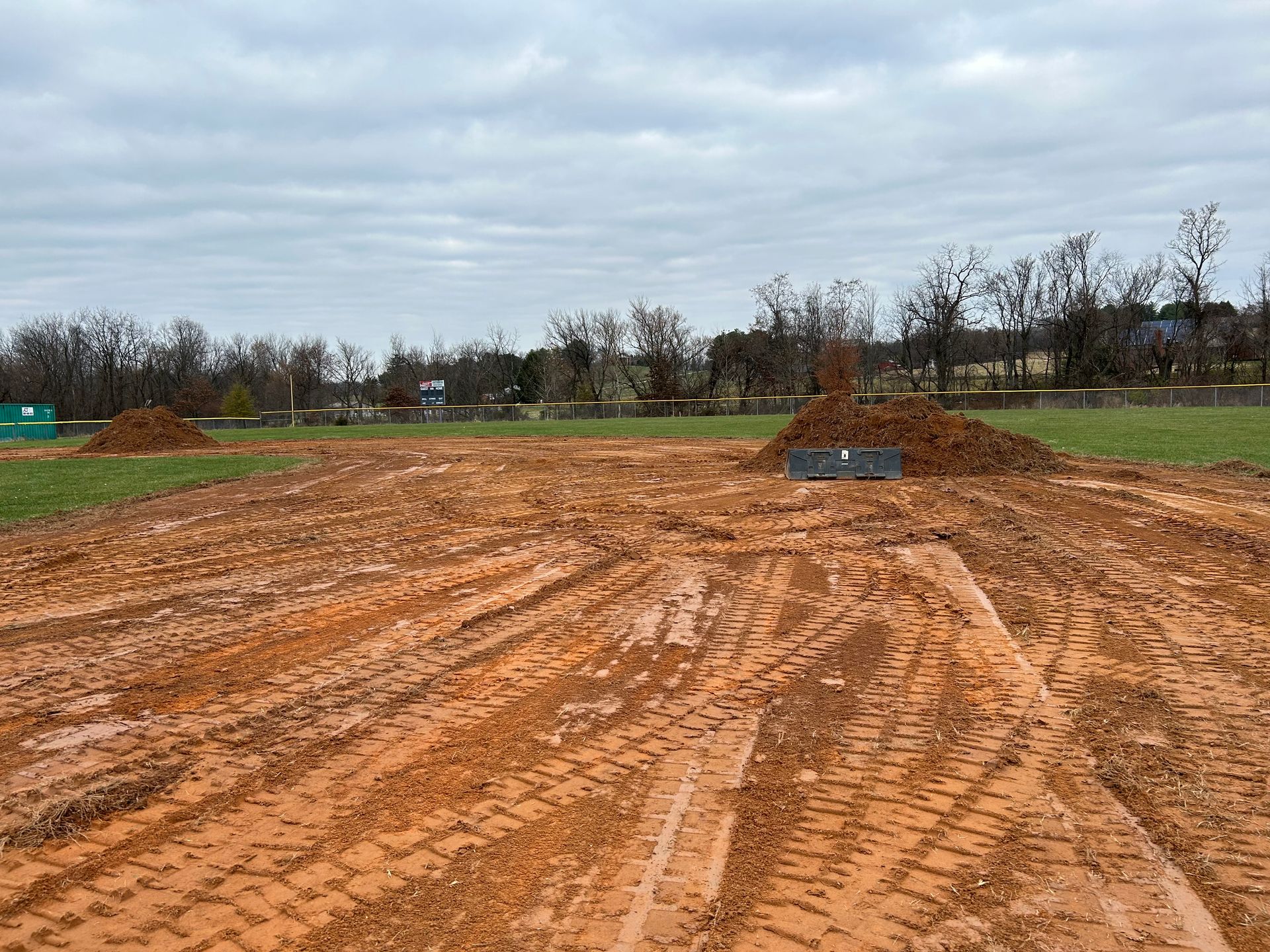 A construction site with reddish-brown dirt, deep tire tracks, and piles of soil under a cloudy, overcast sky.