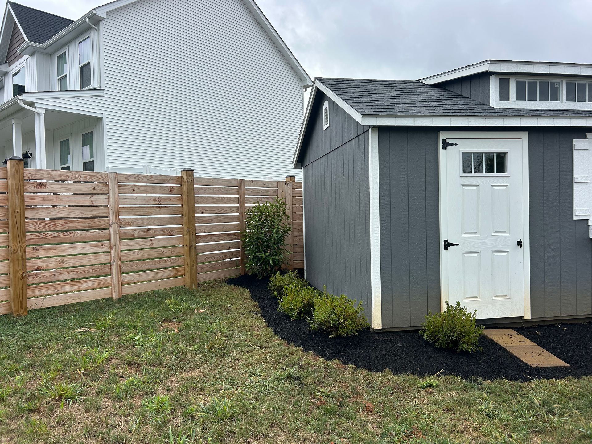 A dark gray shed with a white door sits in a yard next to a horizontal wood fence and a house with white siding.