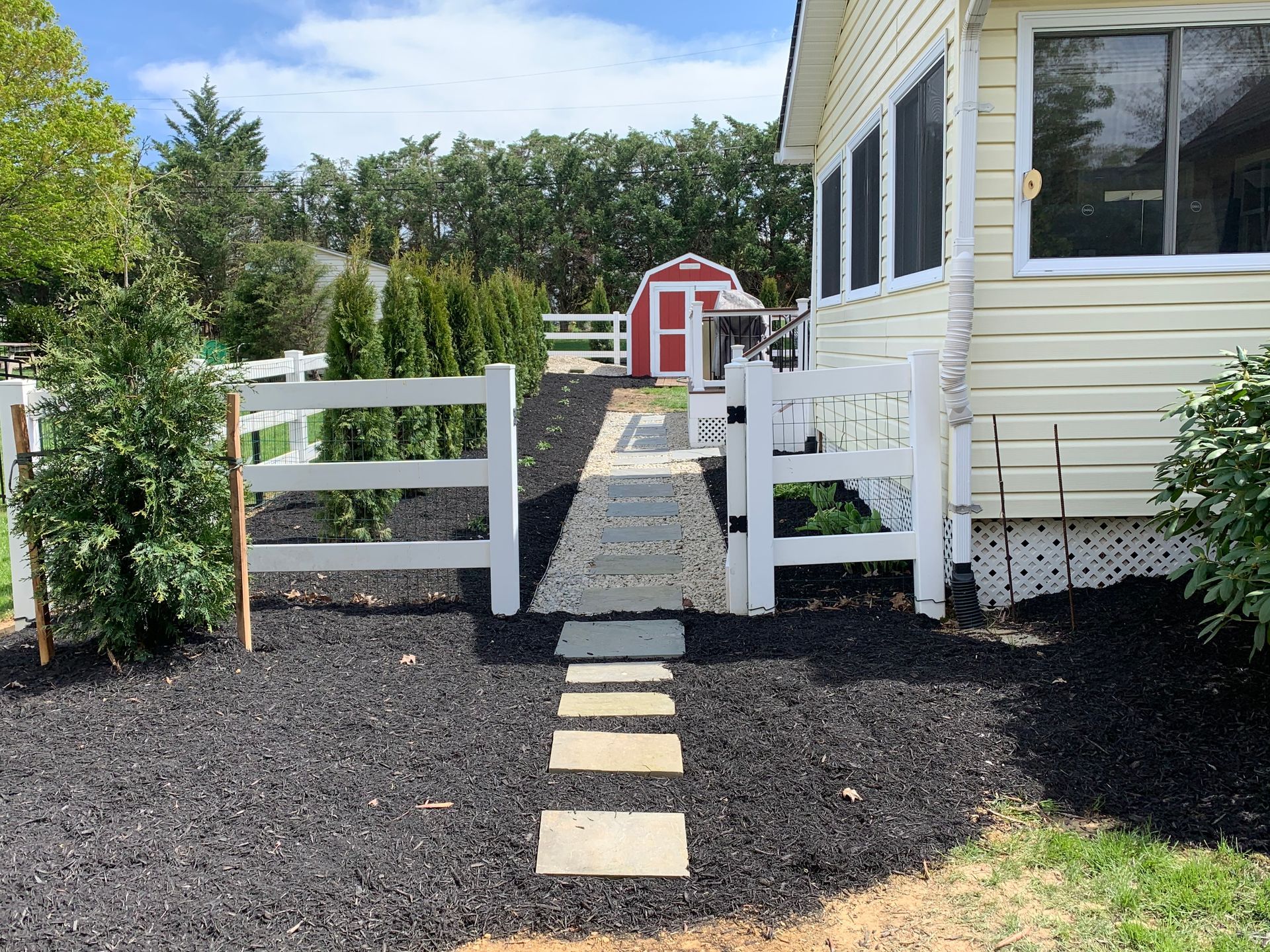 A white fence opens to a stone path leading to a red shed beside a yellow house, all surrounded by black mulch.