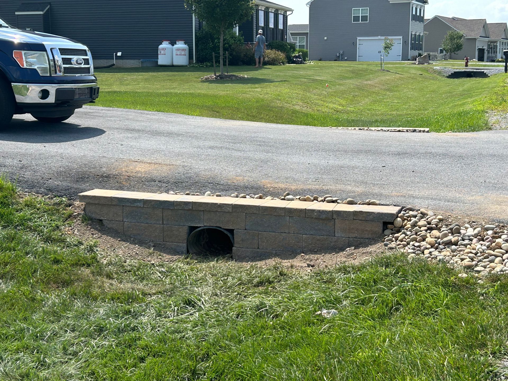 A stone-block retaining wall surrounds a drainage pipe at the edge of a gravel driveway next to a grassy yard.