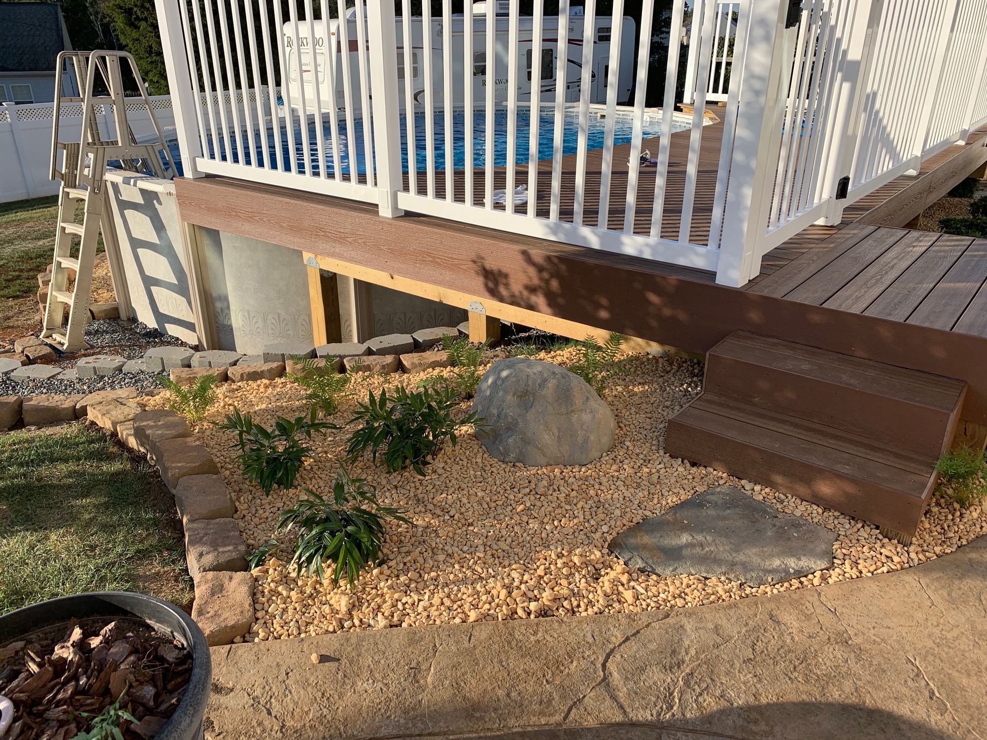 A backyard area with a white-railed deck, gravel landscaping featuring small shrubs and rocks, and stone steps.