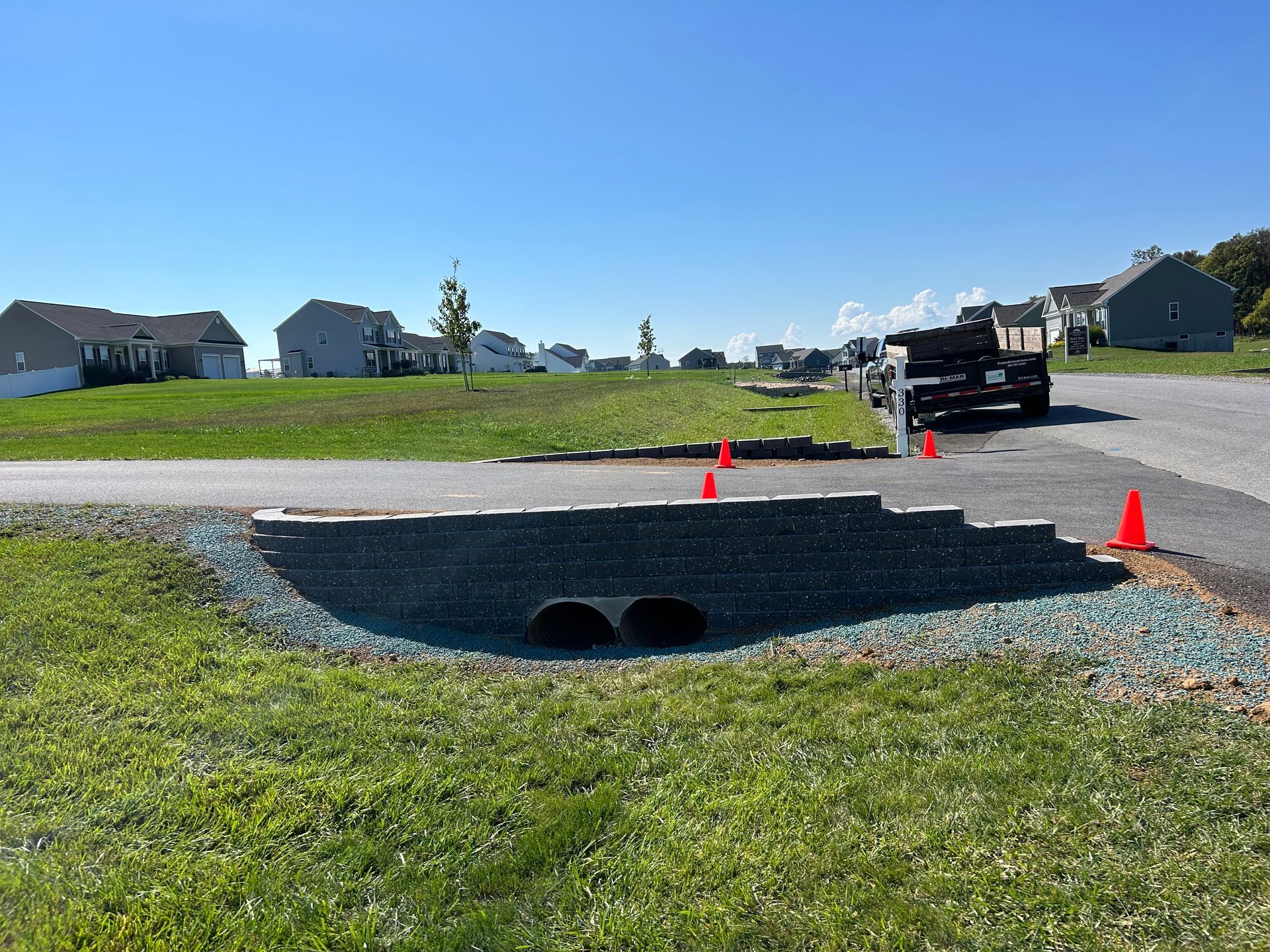 A gray stone retaining wall with a culvert entrance borders a paved driveway in a residential suburban neighborhood.