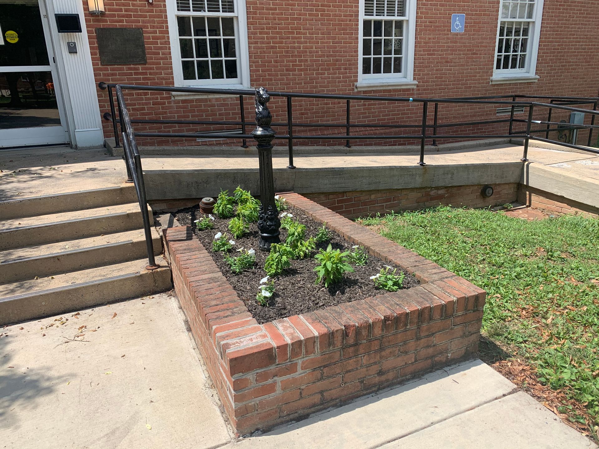 A brick planter with small green plants and a central black post, situated by brick building stairs and a metal railing.