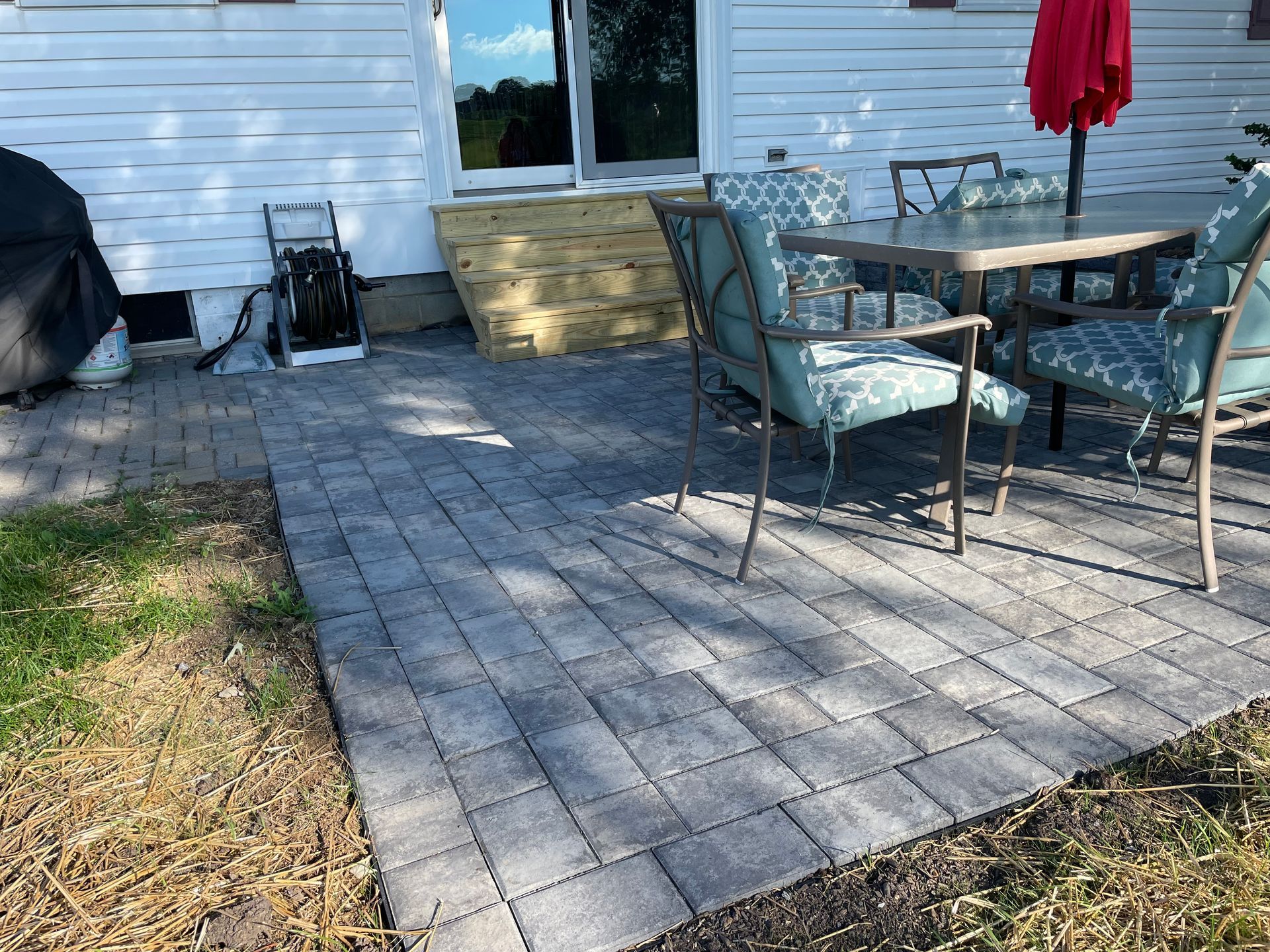 A patio with gray stone pavers, a small wooden staircase, and a table set with chairs beside a white-sided house.
