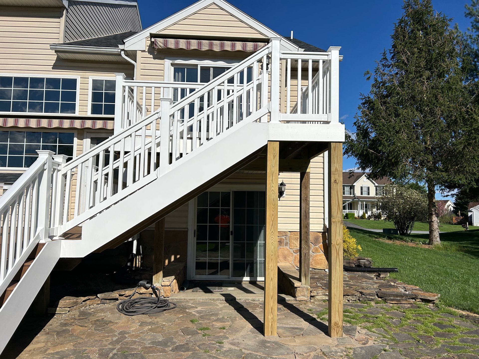 A white wooden staircase with railings leads up to a deck attached to the back of a tan house with a stone patio below.