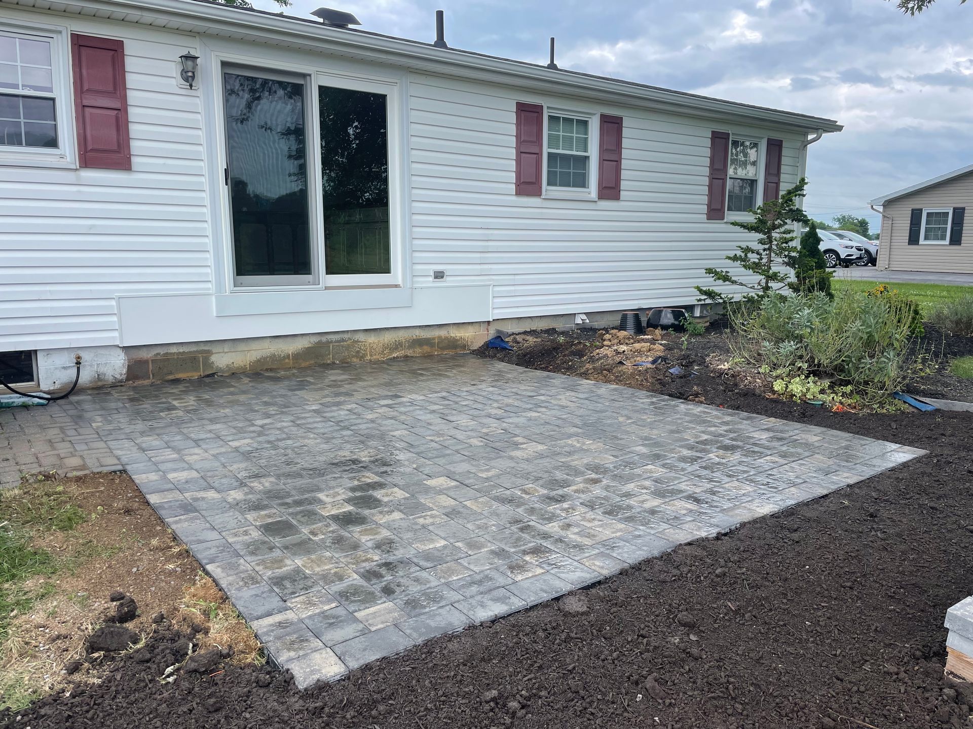 A newly installed gray paver patio sits outside a white-sided house with red shutters and a sliding glass door.