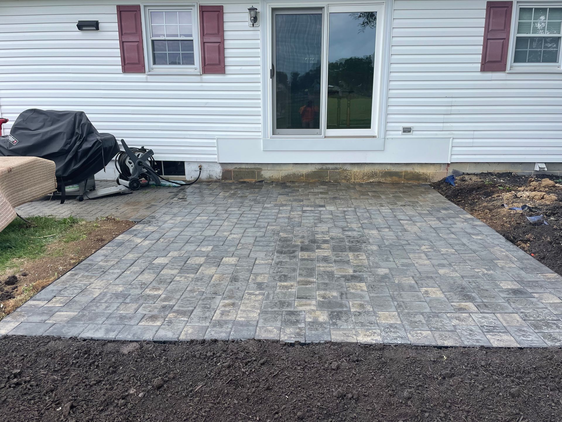 A newly installed gray paver patio sits in front of a house with white siding and a sliding glass door.