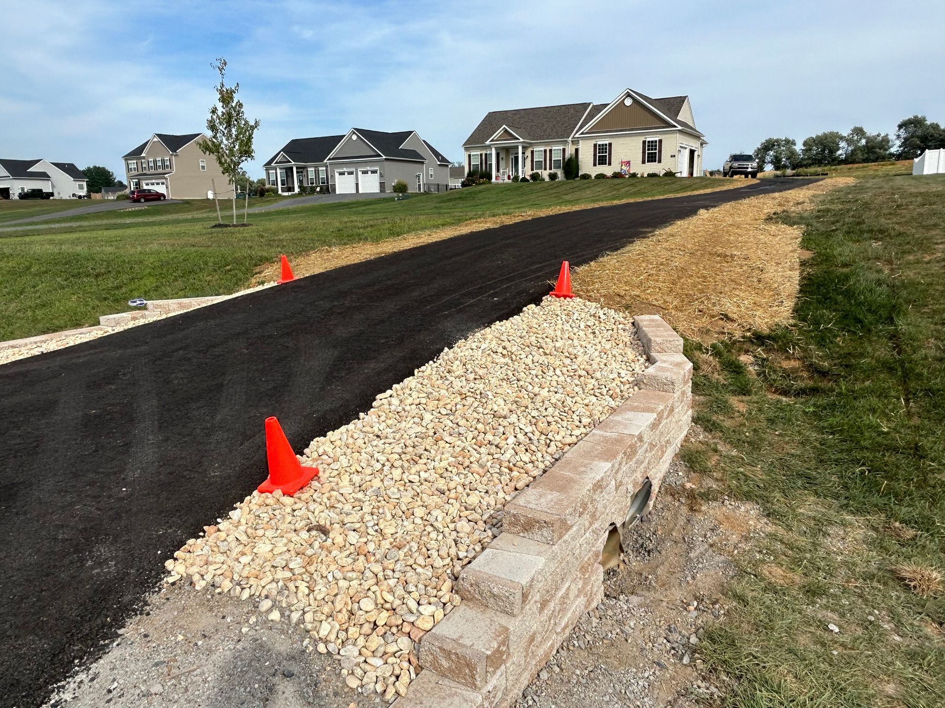 A newly paved asphalt driveway leads to houses, featuring a retaining wall, gravel border, and orange traffic cones.
