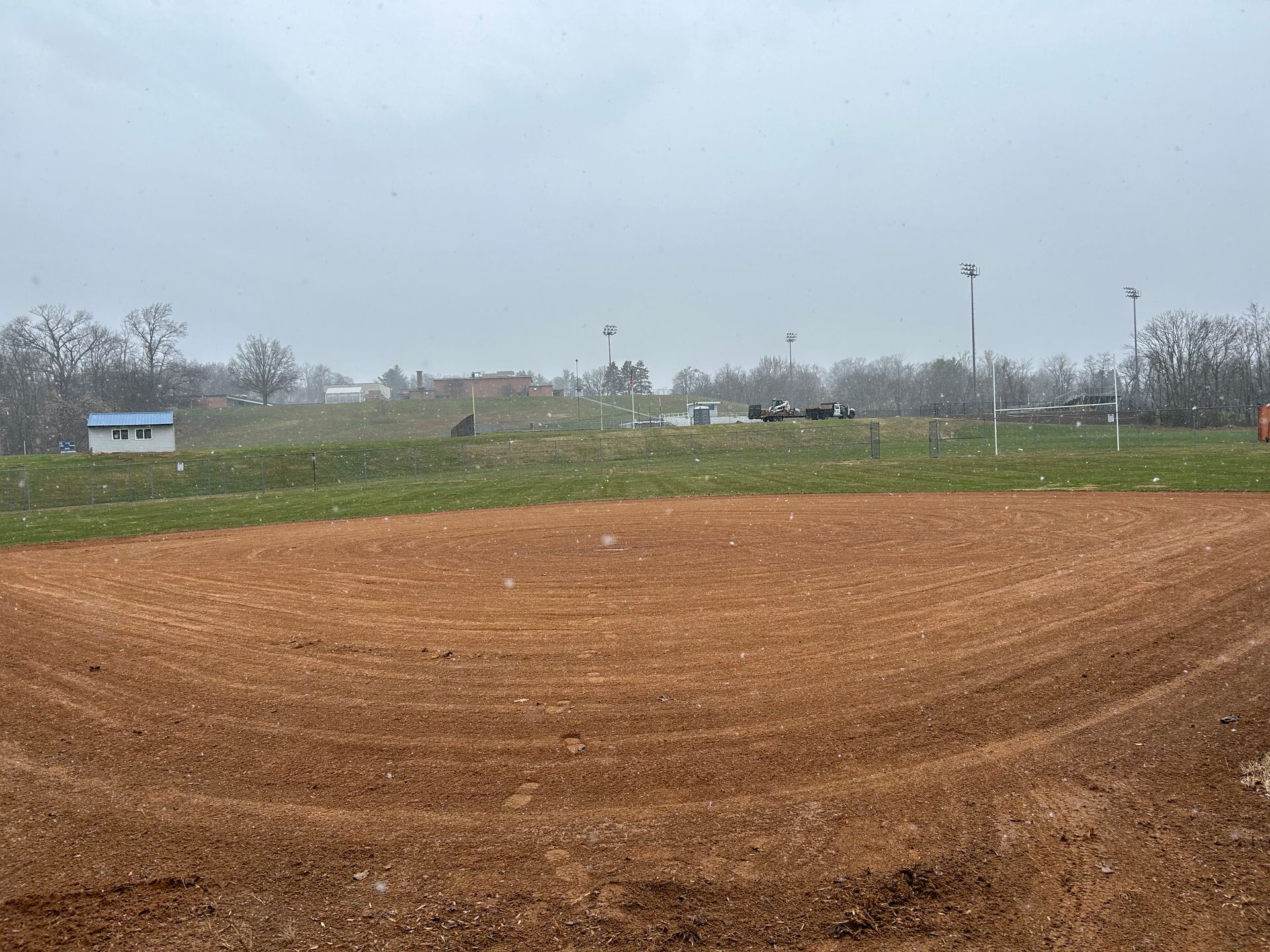 A dirt baseball infield on a cloudy day, with a small building and light poles visible in the background.