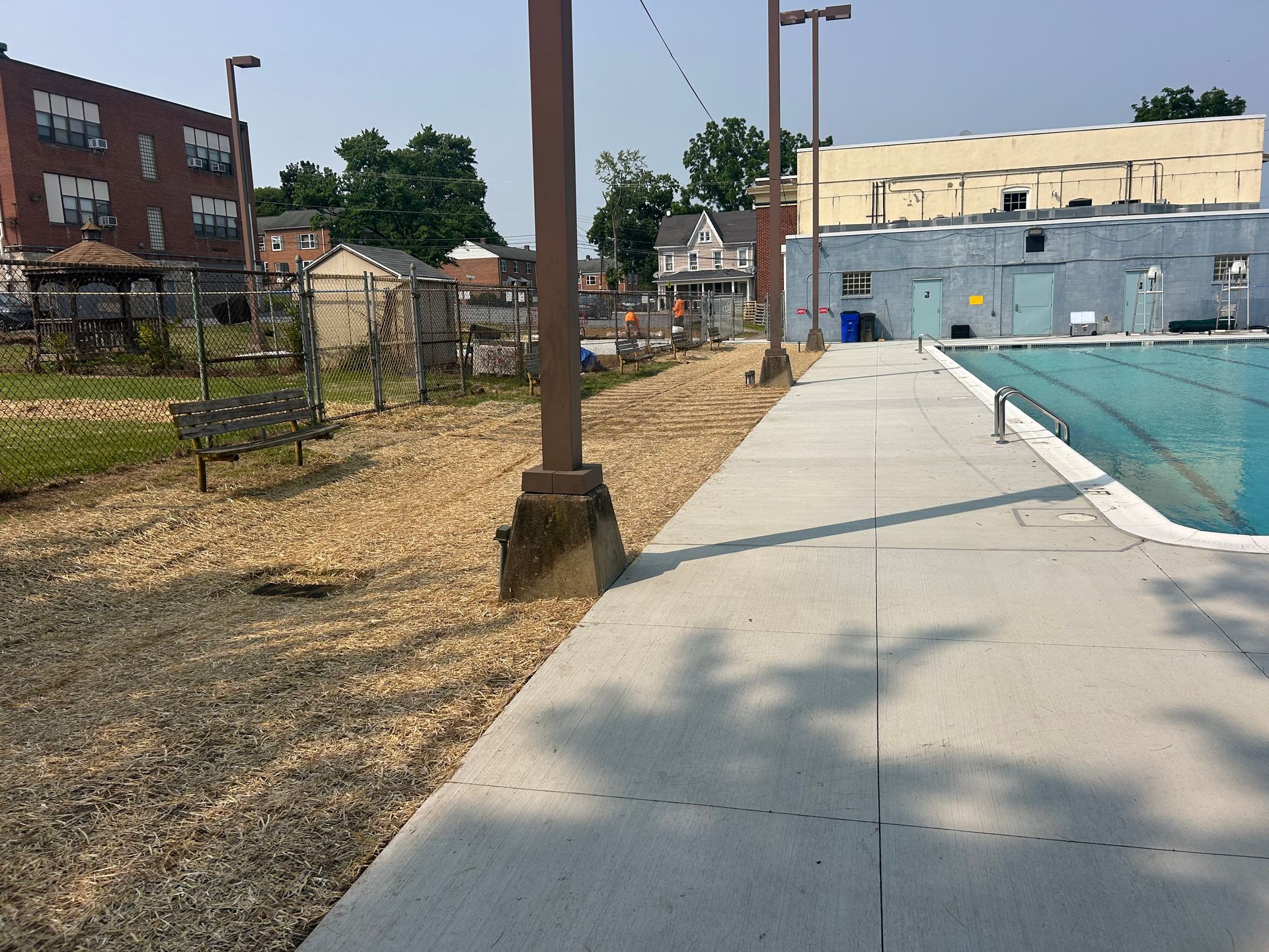 A concrete pool deck runs alongside a swimming pool, adjacent to a dirt lot with a bench, shed, and surrounding buildings.