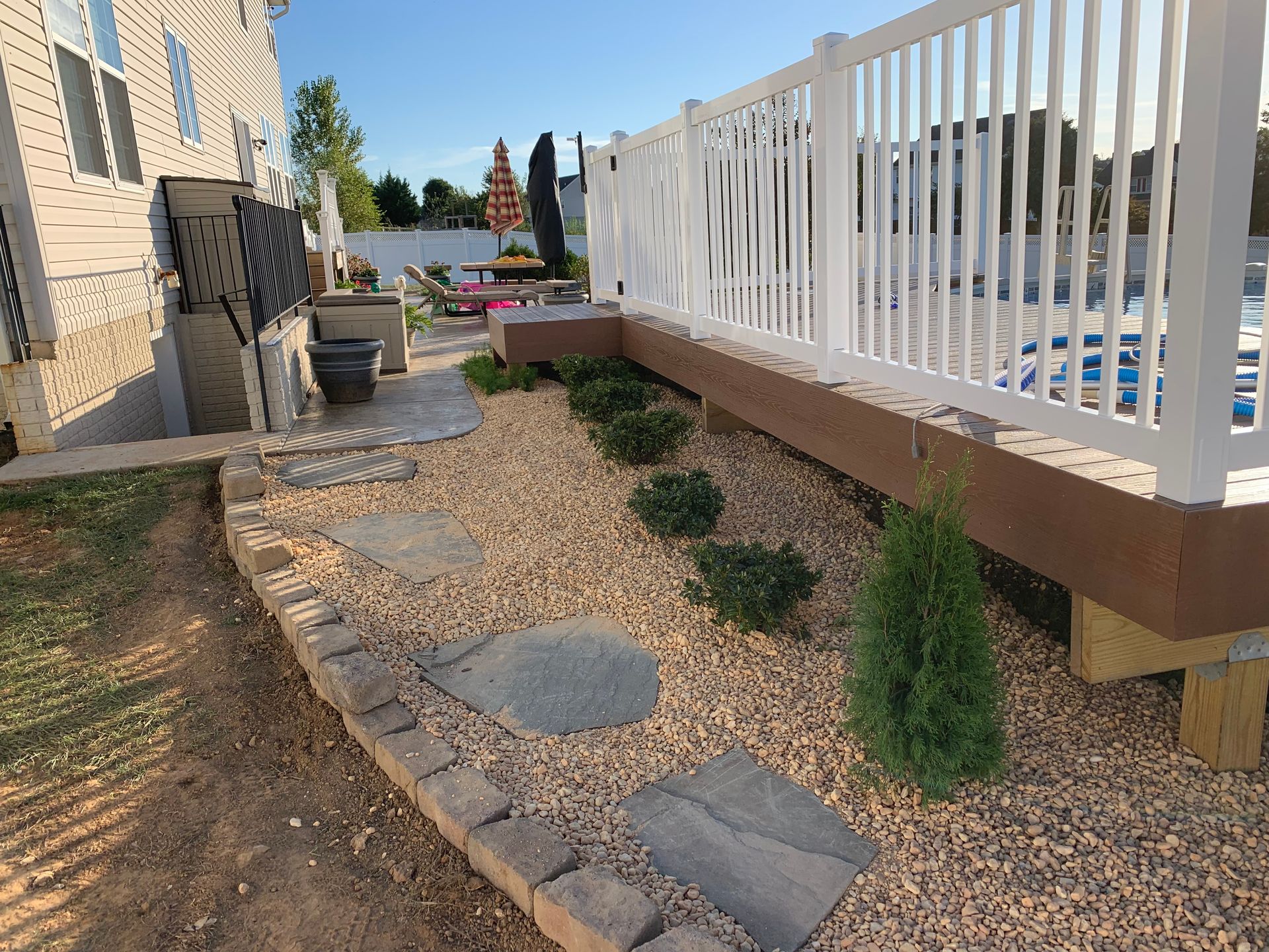 A stone path winds through a gravel landscape bed next to a raised white deck with a pool in a residential backyard.