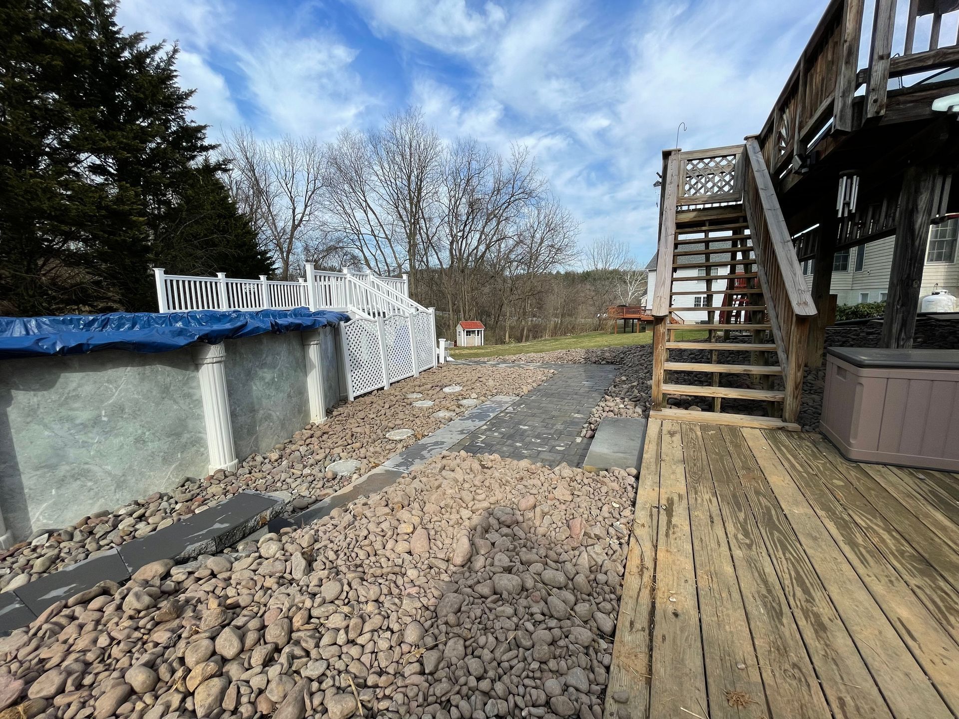 A backyard featuring a pool covered with a blue tarp, a wooden deck with stairs, and a ground covered in brown gravel.