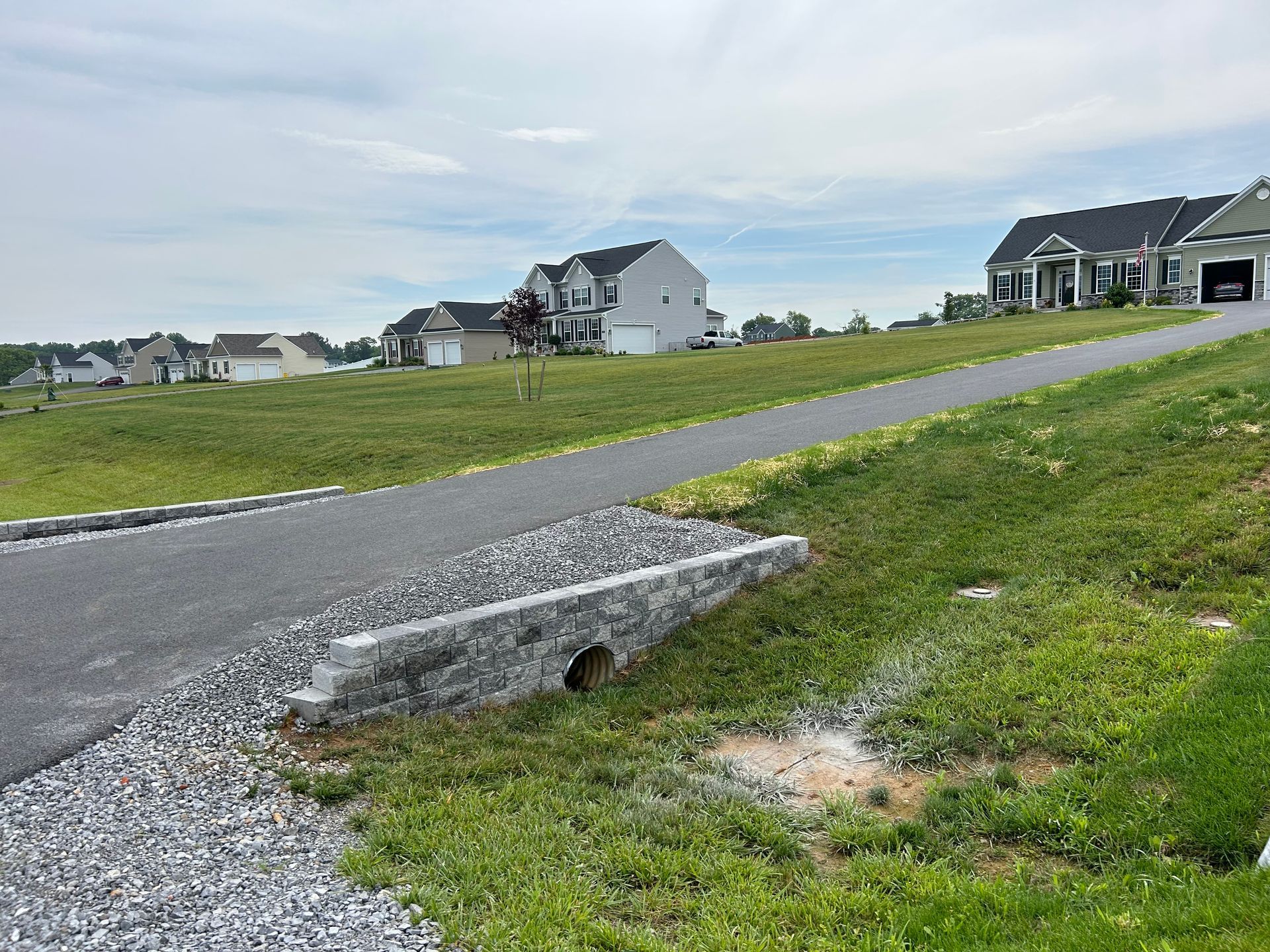 A paved path crosses a culvert with a stone retaining wall in a grassy residential neighborhood under a cloudy sky.