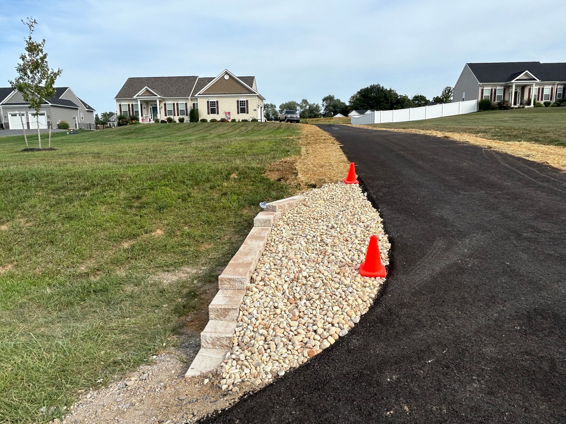 A stone retaining wall bordering an asphalt driveway with gravel filler and two orange traffic cones in a grassy yard.