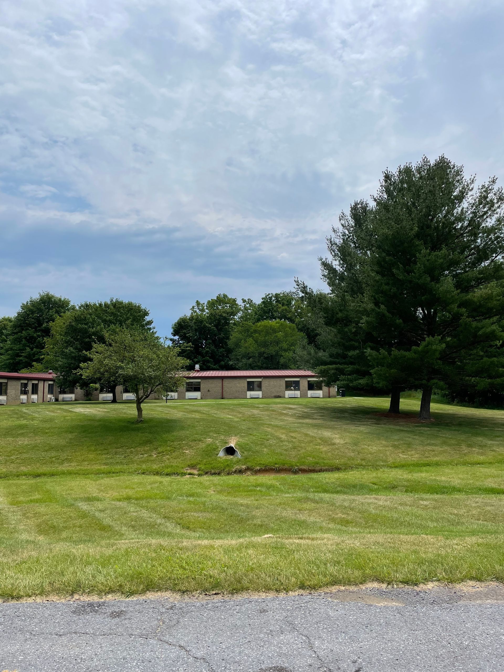 A one-story building with a red roof sits on a grassy hill behind trees, under a cloudy sky.