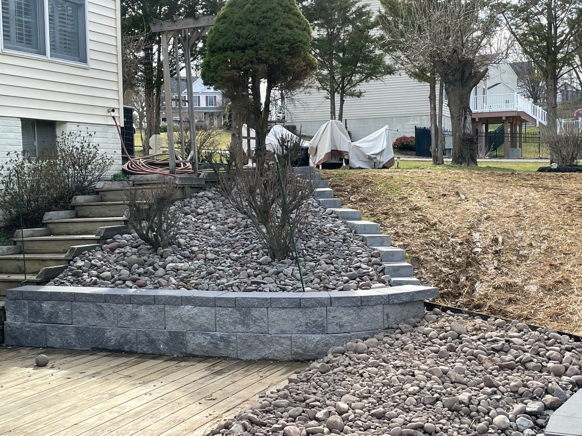 A tiered landscape featuring gray stone retaining walls, rock-filled garden beds, stairs, and evergreen shrubs.