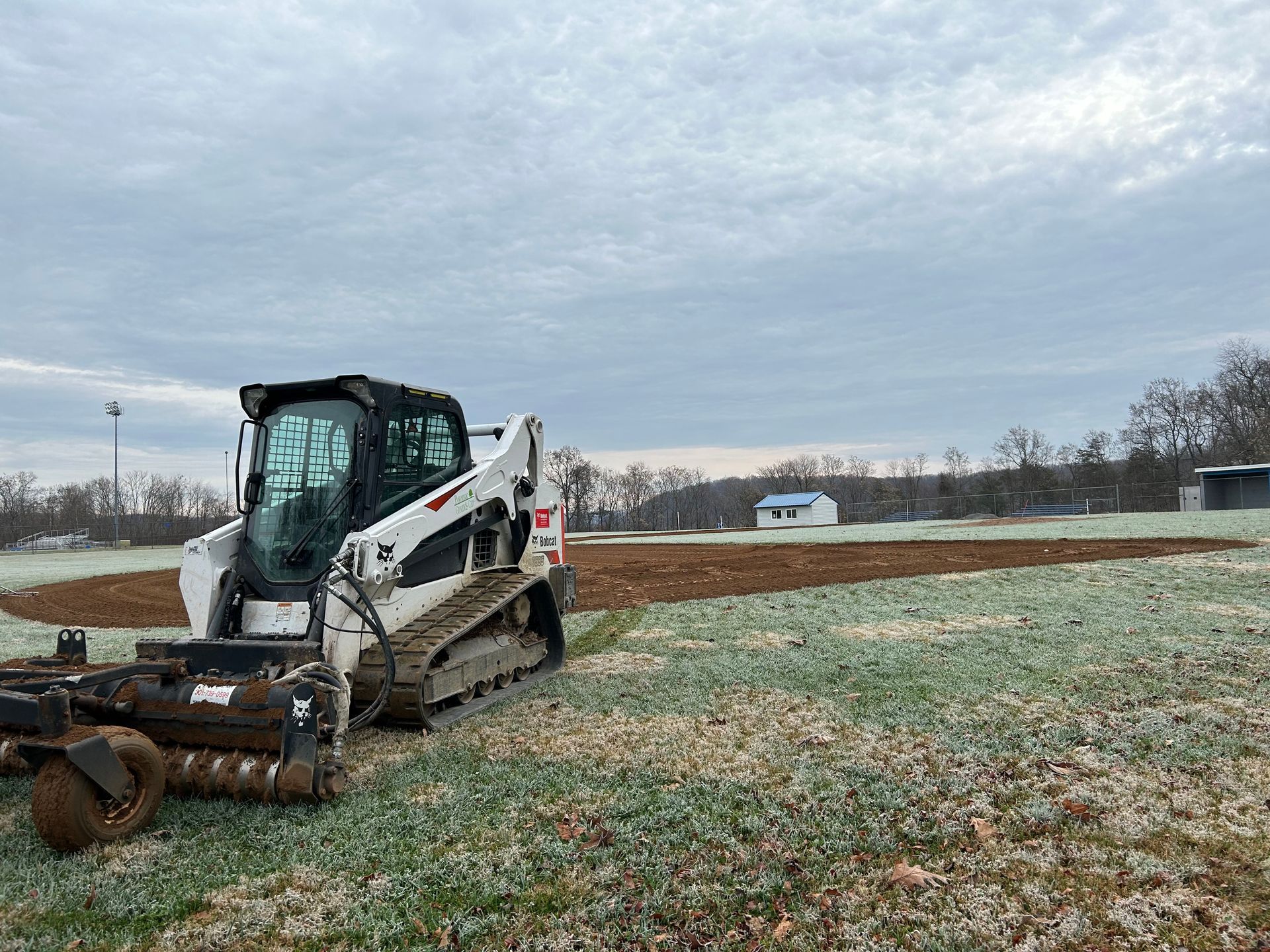 A white Bobcat compact track loader with a mower attachment sits on a frost-covered field next to a tilled patch of earth.