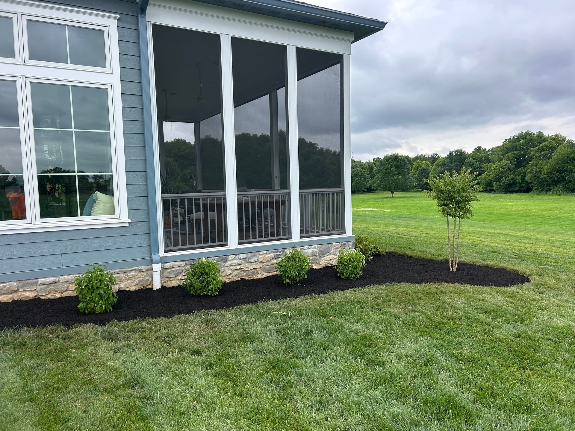 A side view of a blue house with a screened-in porch and stone foundation, featuring a landscaped mulch bed with shrubs.