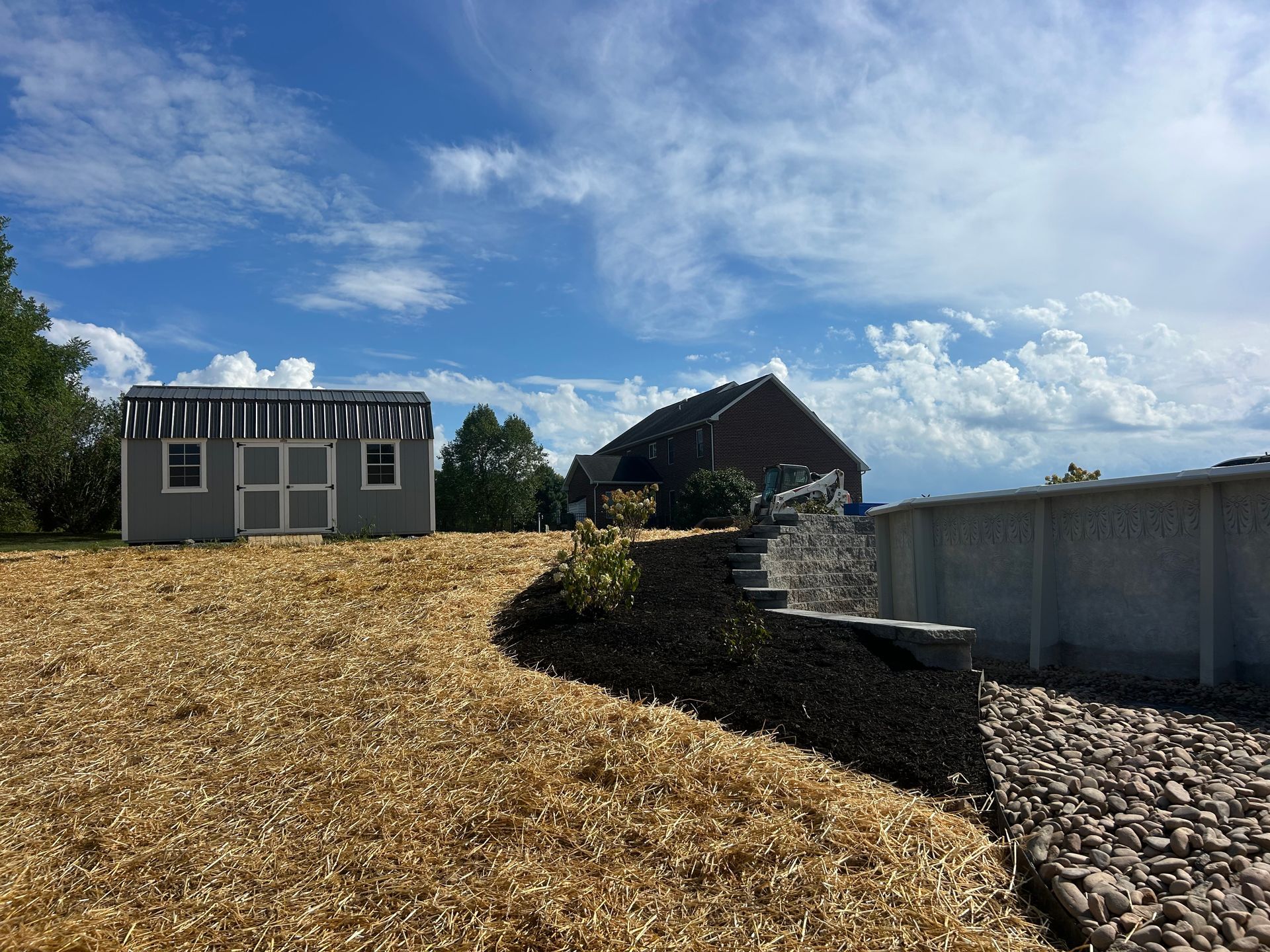 A gray storage shed sits on a mulch-covered yard beside a stacked stone retaining wall and an above-ground pool.