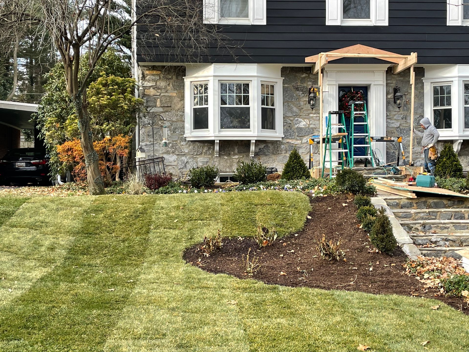 A person stands on the stone steps of a home with a dark exterior, a stone facade, and a newly landscaped front yard.