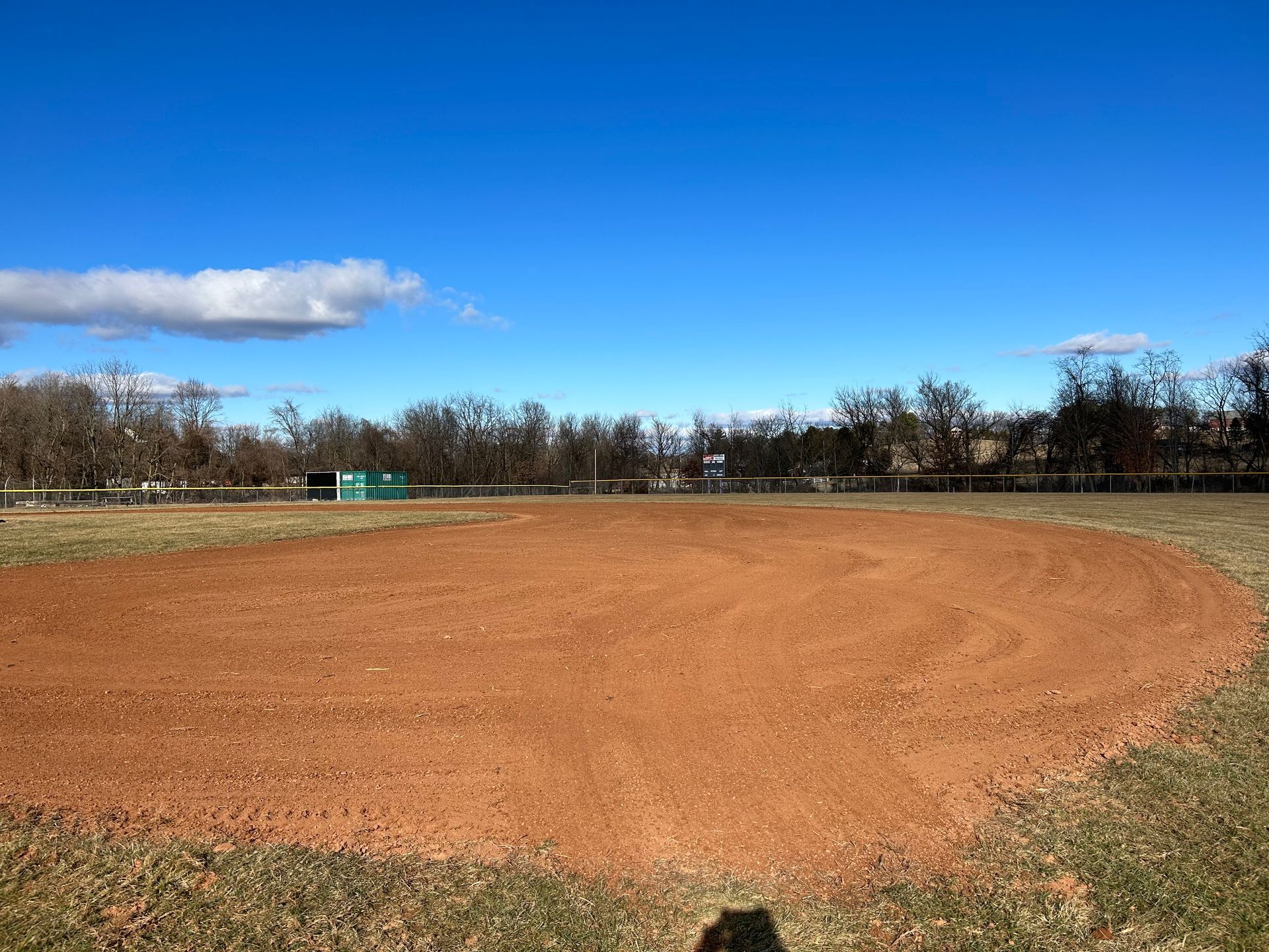 A patch of reddish-brown dirt forming a baseball infield against a backdrop of bare trees under a clear blue sky.