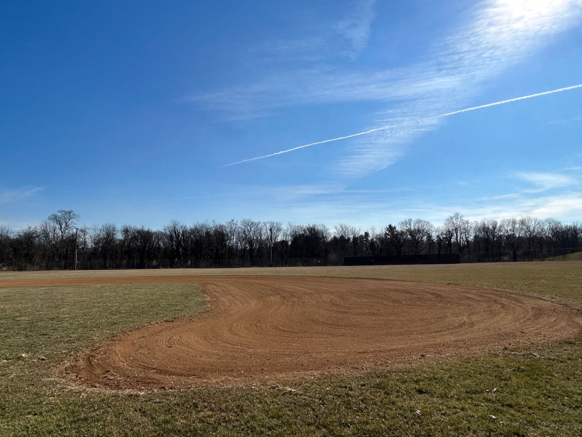 A dirt baseball infield cutout sits in a grassy field under a bright blue sky with a wispy airplane contrail.