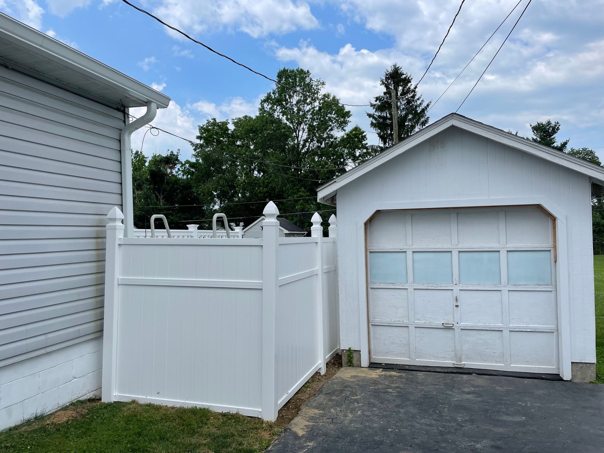 White vinyl privacy fence connecting the corner of a house to a white detached garage on a paved driveway.