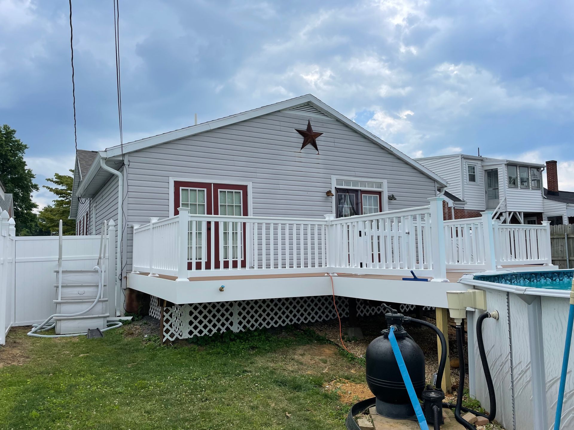 A gray house with a white deck, red doors, and a decorative brown star, beside an above-ground pool in a grassy yard.