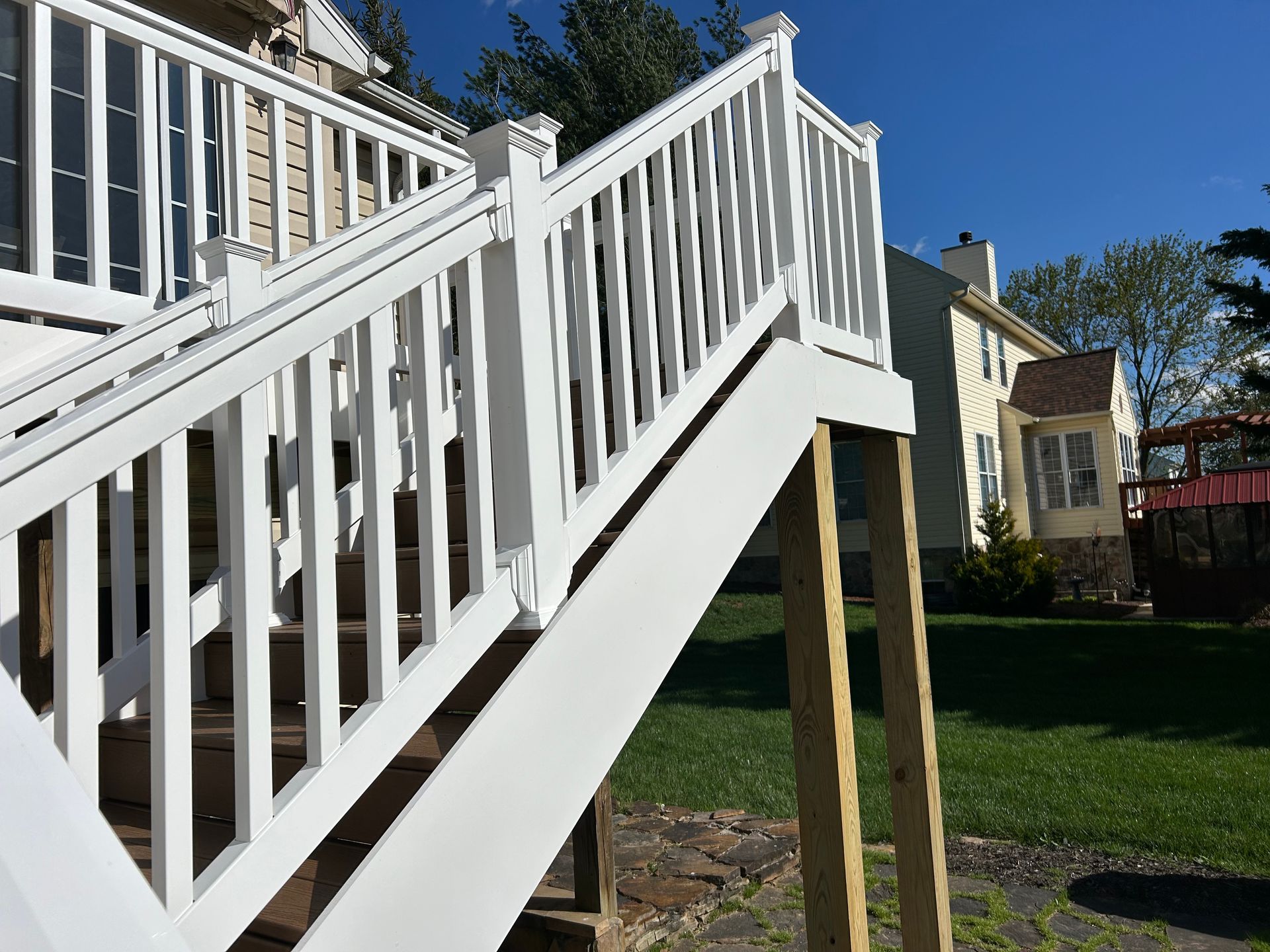 A white wooden staircase with railings leading up to a house deck on a sunny day with a green lawn in the background.
