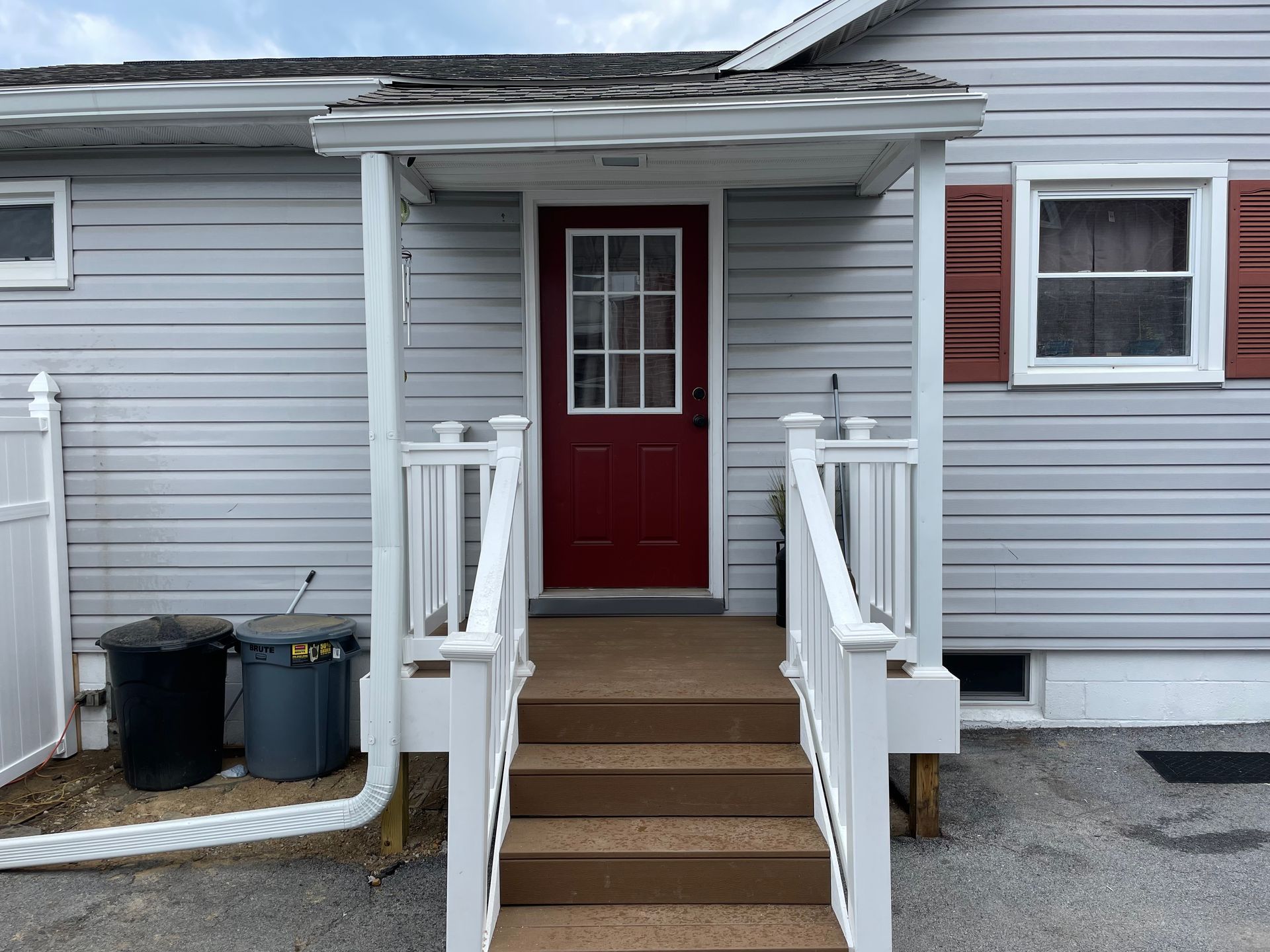 A gray house with a red front door, a small porch with white railings, and two trash cans on the side.