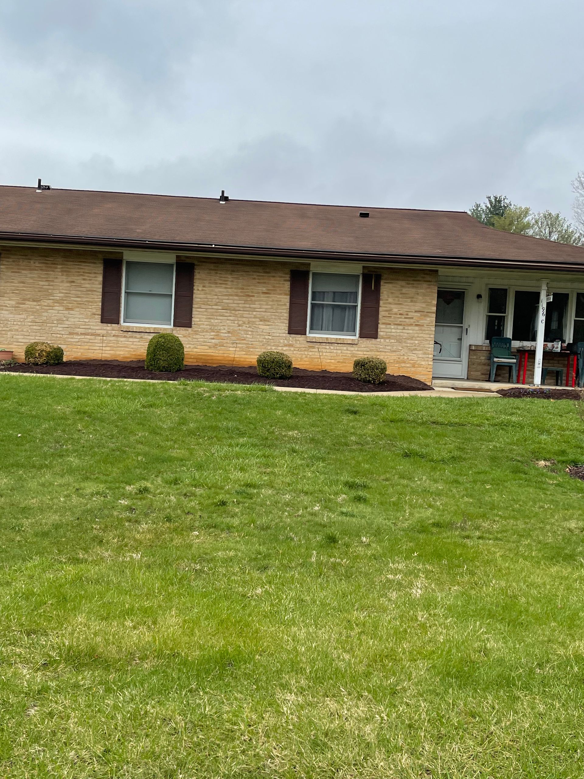A single-story house with tan stone siding, dark brown shutters, and a brown shingled roof, set behind a green lawn.