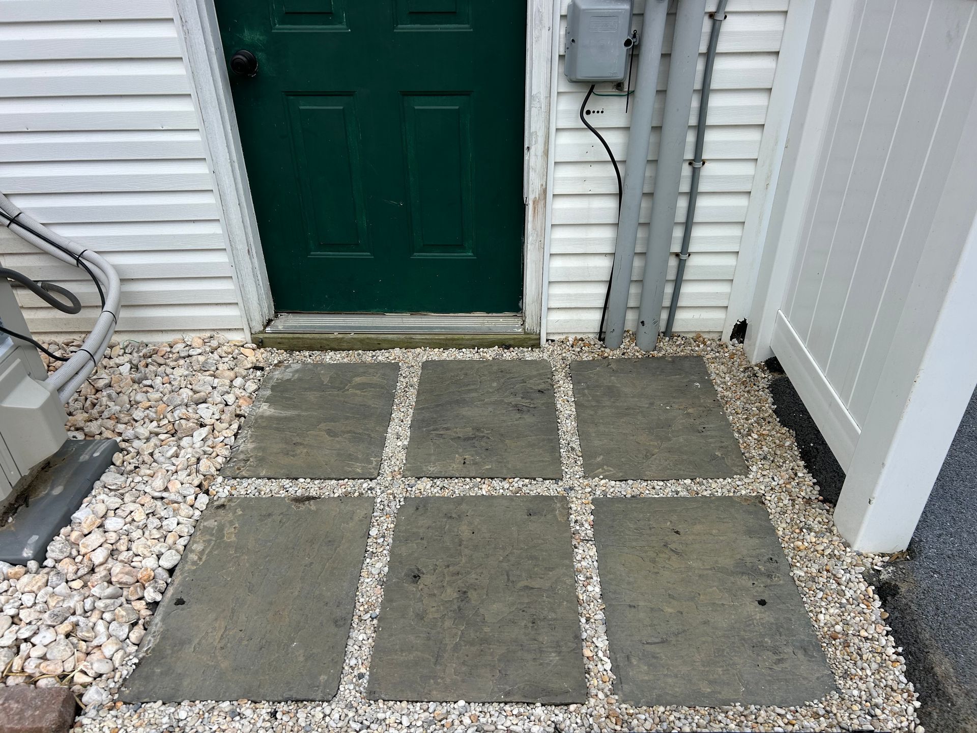 A small entryway with a green door, featuring six flat rectangular gray pavers surrounded by white gravel.