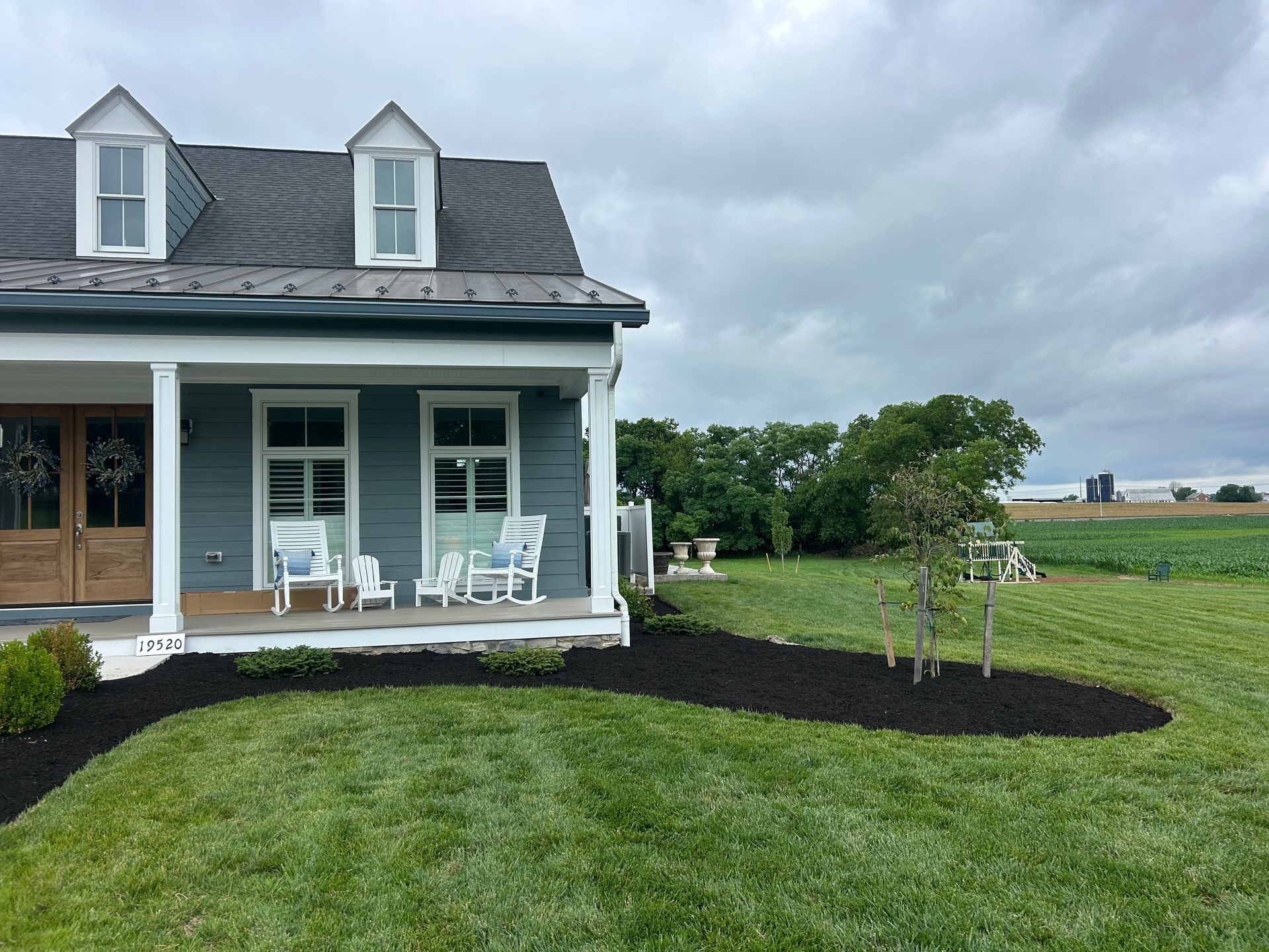 A gray house with a front porch, white rocking chairs, and fresh mulch landscaping under a cloudy sky near a field.