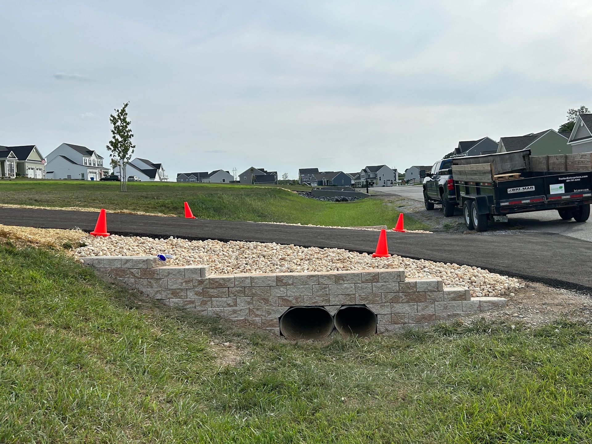 A stone retaining wall built over two culverts beneath an asphalt path, with traffic cones and a trailer parked nearby.