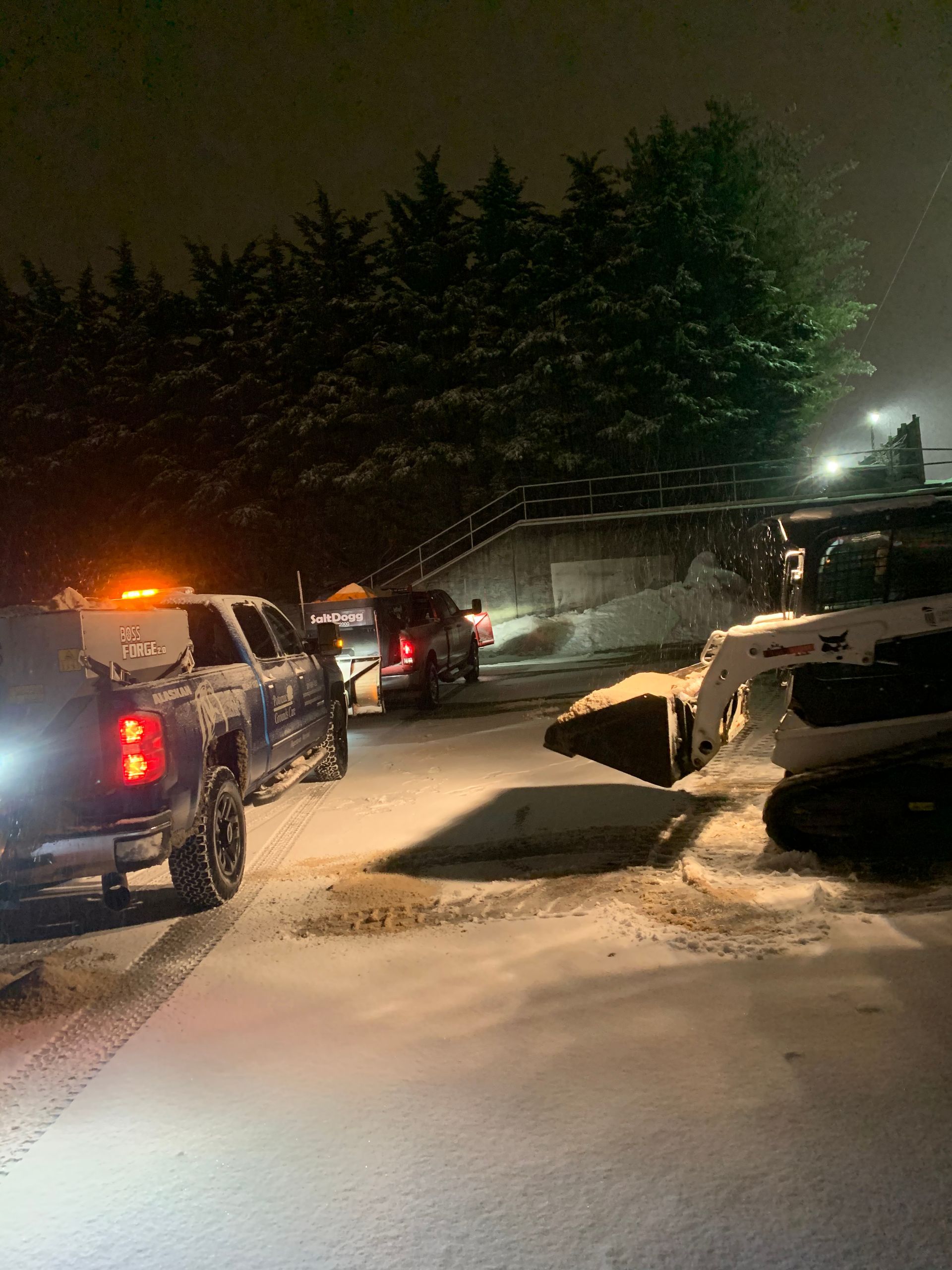 A nighttime scene shows a skid-steer loader and pickup trucks parked in a snowy parking lot near a tree line.